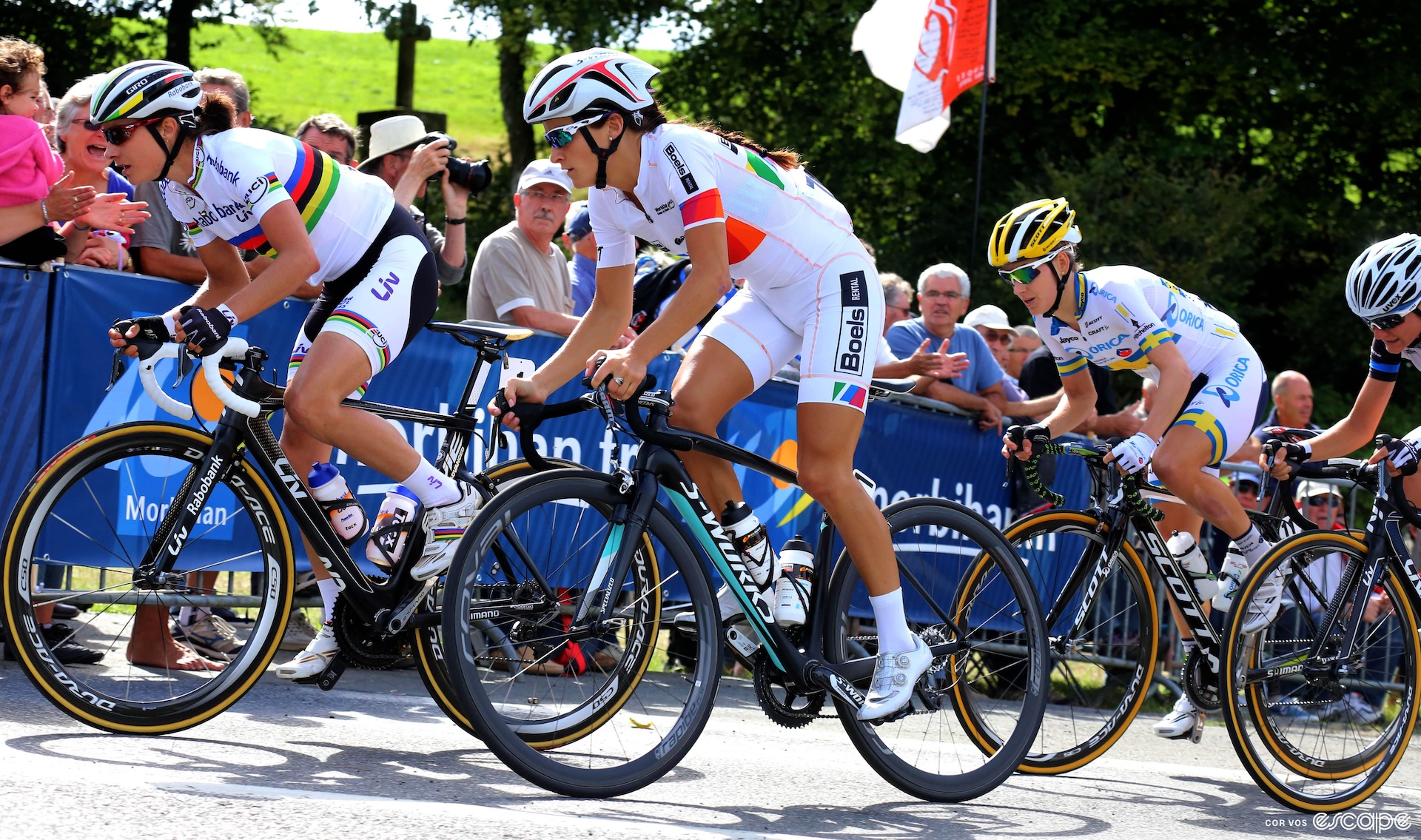 A group of women race bikes up a hill, one wears white bib shorts