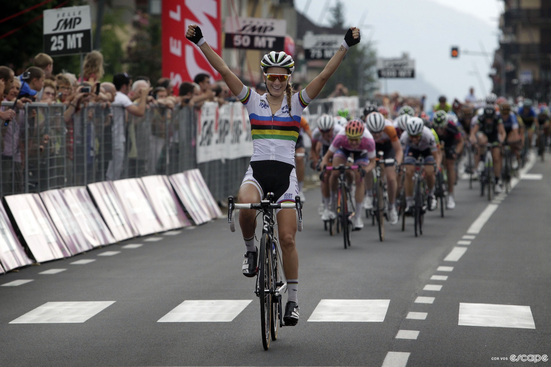 A woman raises her arms in victory as she wins a bike race, behind a group of women on bikes chases her.