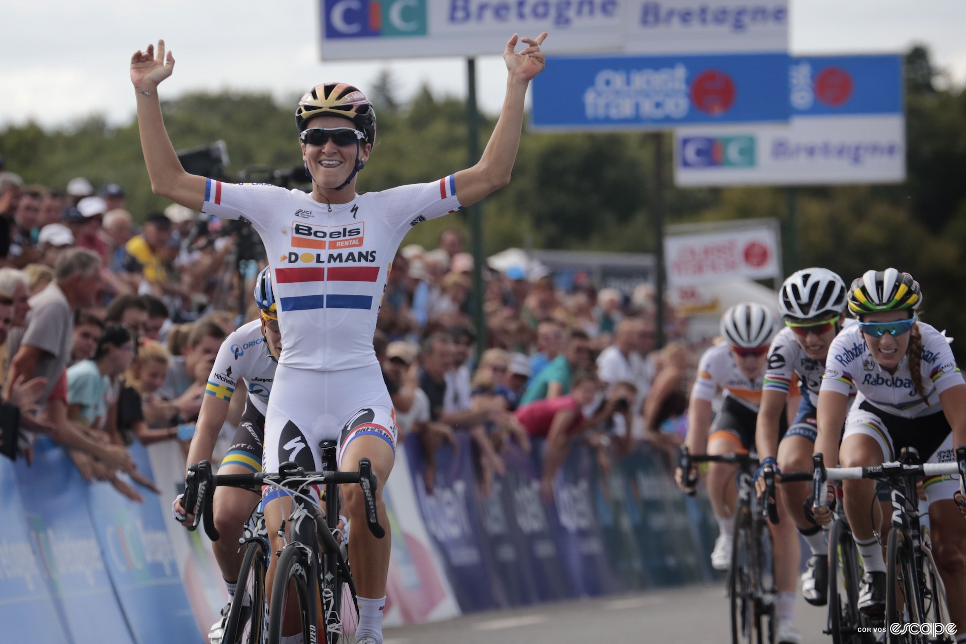 A cyclist in all white throws her arms in the air as she wins a bike race
