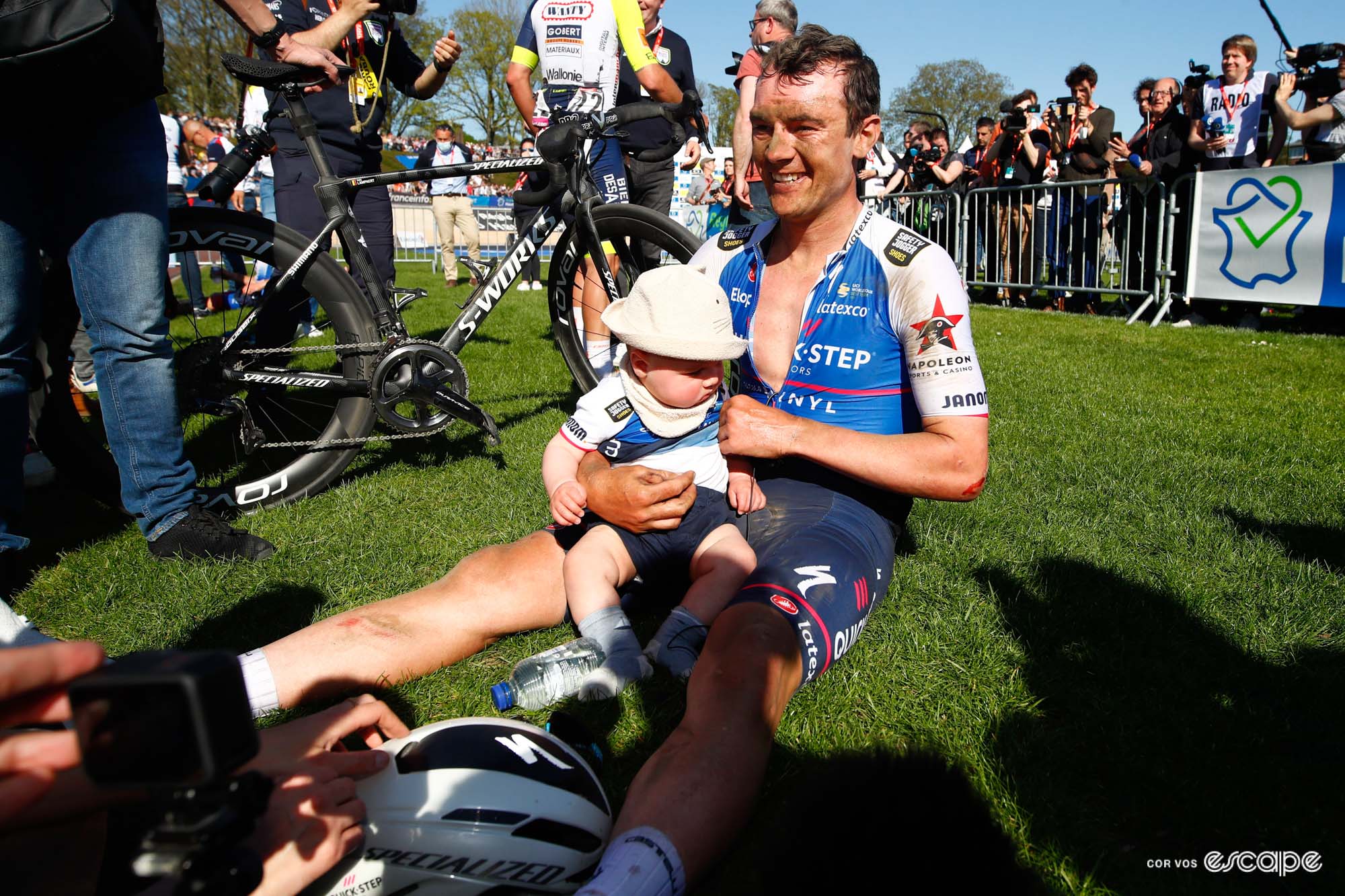 Yves Lampaert sits on the green infield grass at the velodrome following the 2022 Paris-Roubaix. His infant son sits on his lap. wearing a team jersey and bucket hat. Yves is smiling and covered with grit.