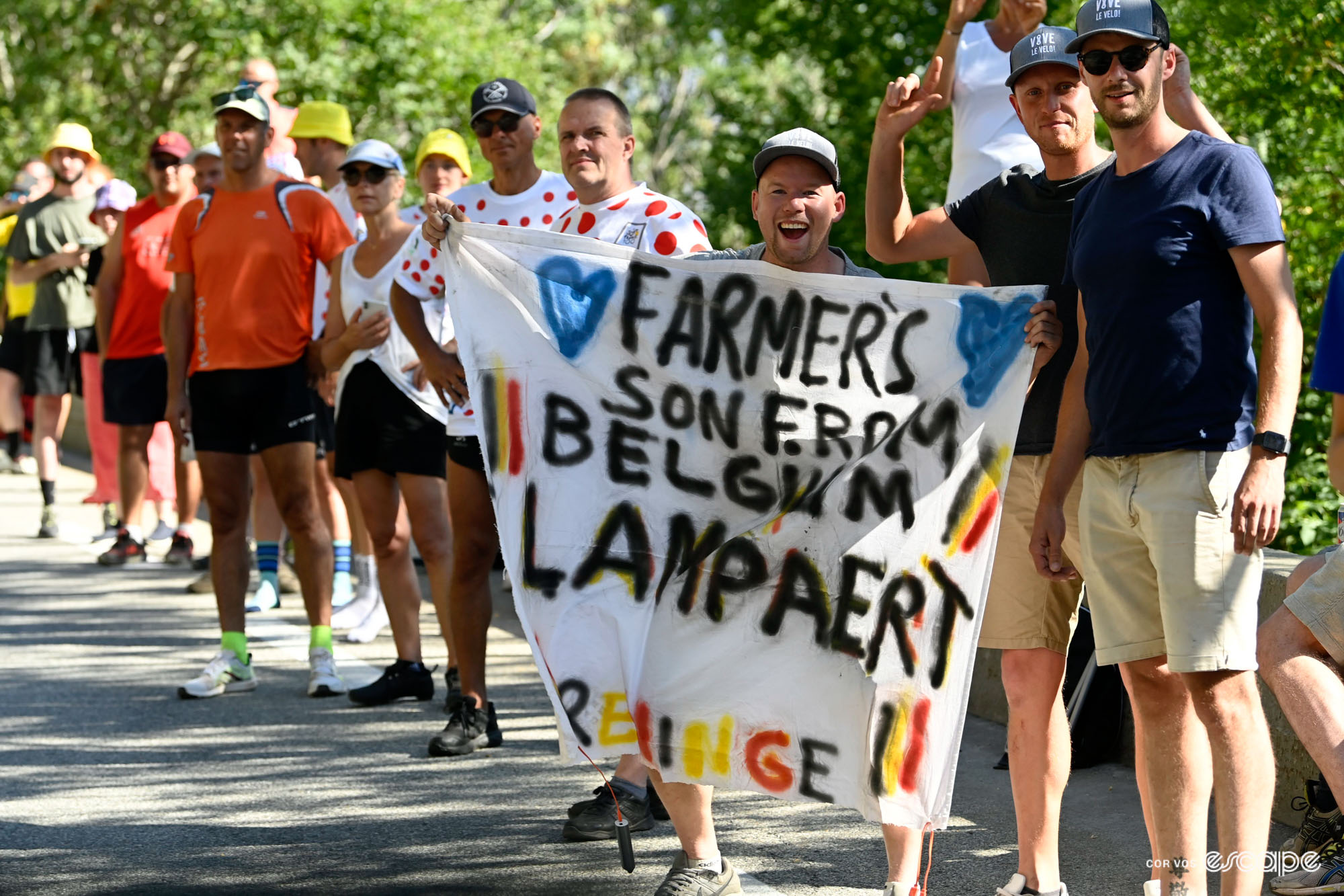 Fans stand with signs on the roadside to cheer Yves Lampaert. This one reads "Farmer's son from Belgium" with blue hearts and belgian flags.