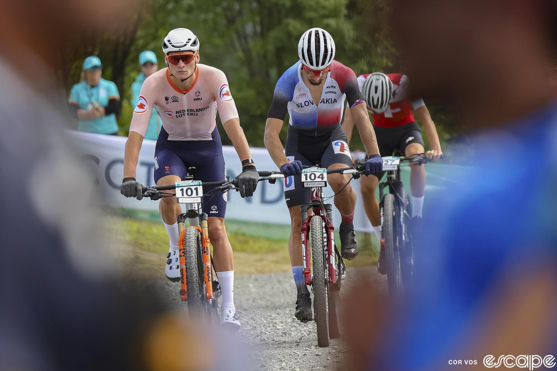 Mathieu van der Poel and Peter Sagan are framed between two blurred out fans as they ride to the start of the 2023 World Mountain Bike Championships in Glasgow, Scotland. They're both in national team kit. Van der Poel looks ahead with a neutral expression, while Sagan is looking down at his computer.