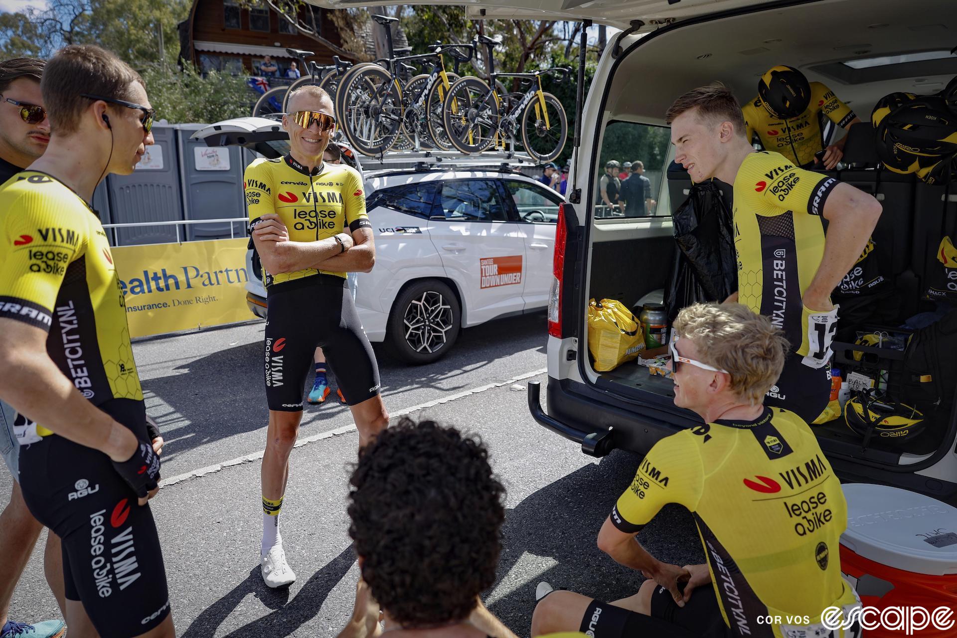 Bart Lemmen, Robert Gesink, and other Visma teammates stand at a team van for a debrief before a Tour Down Under stage. They look relaxed, and Gesink, wearing wraparound sunglasses, is smiling as he appears to hold court.