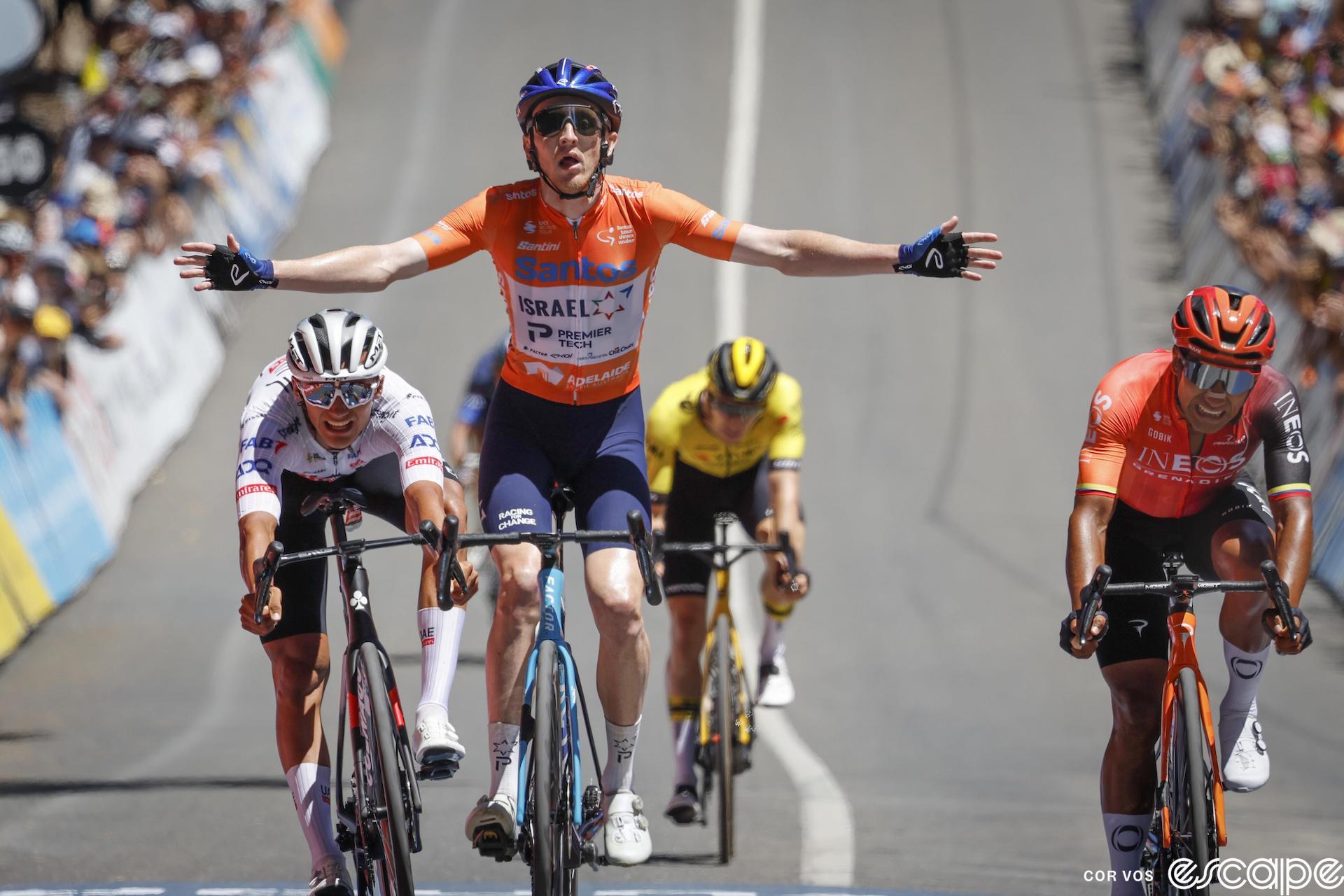 Stephen Williams raises his arms to win the Willunga Hill stage of the 2024 Tour Down Under. Just behind, slightly out of focus, is a yellow-clad Bart Lemmen.