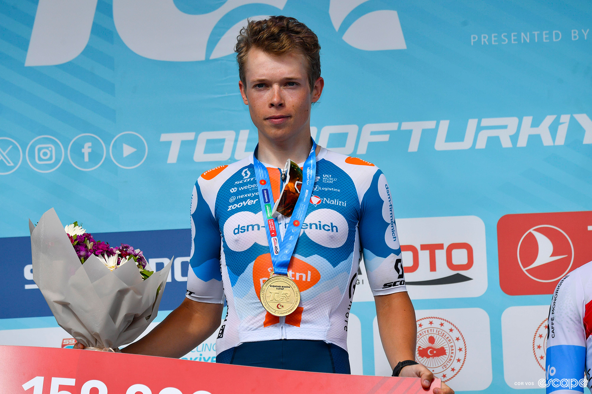DSM Firmenich-PostNL's Frank van den Broek stands on the podium of the Tour of Turkey. He has a large winner's medal around his neck and a bouquet of flowers in his right hand, and is wearing a carefully neutral expression.