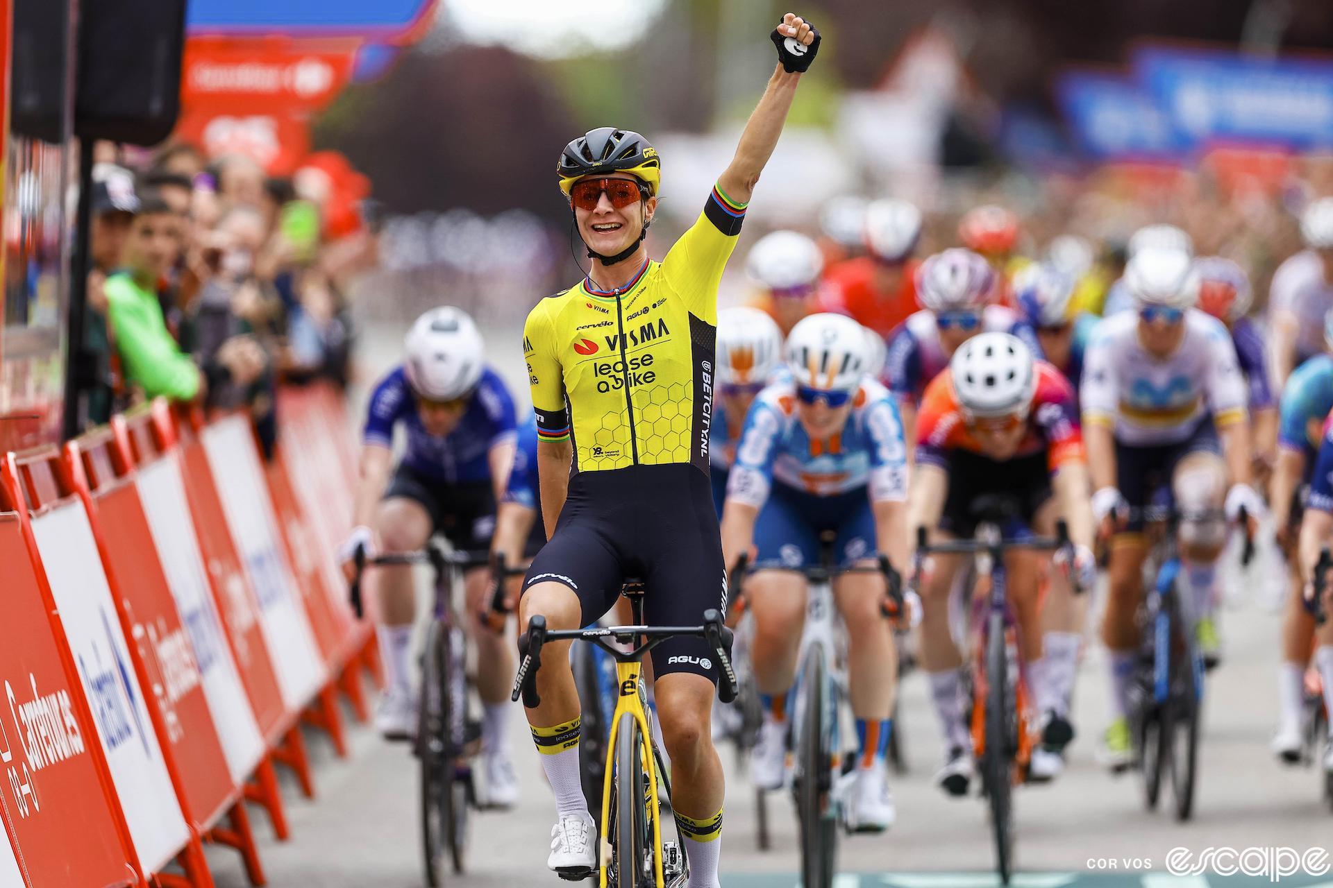 Marianne Vos raises her arm in victory as she leads the peloton across the line to win stage 3 of the Vuelta España Femenina. She's markedly in front of the chasing pack, led by Charlotte Kool.