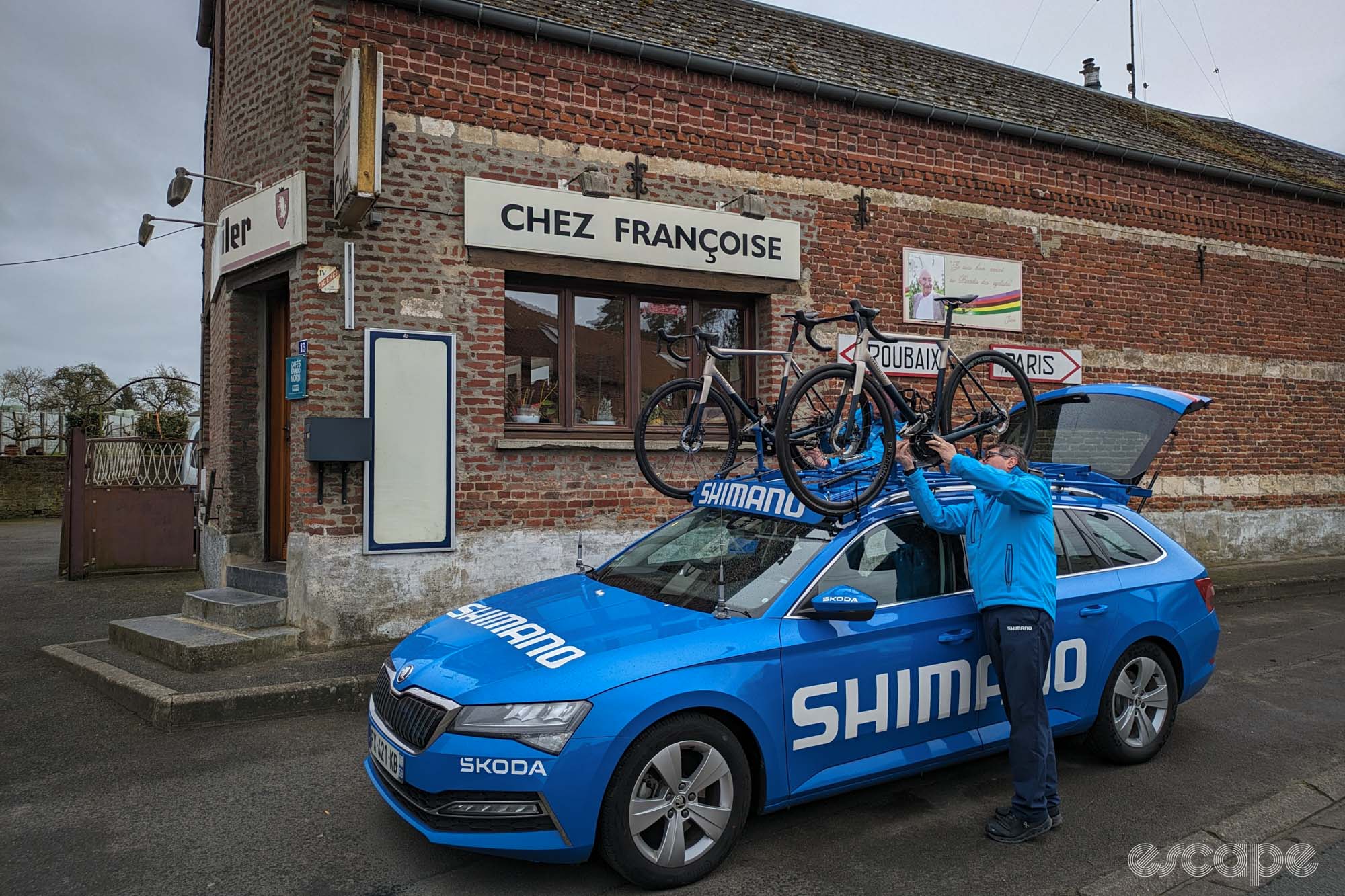 Bruno Mallet checks a spare bike atop the blue Shimano neutral service car outside the brown brick Chez Francoise building.
