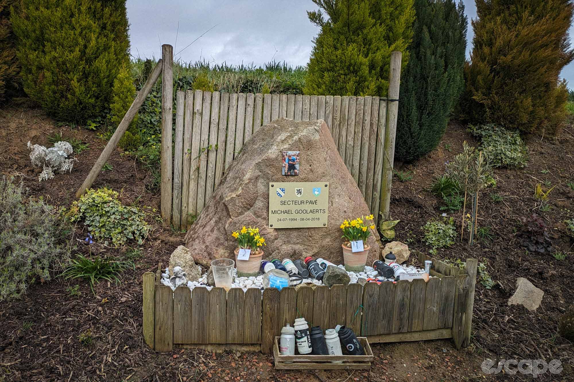 The Michael Goolaerts memorial features a plaque on a large stone, adorned by water bottles left by fans and two pots of plastic daffodil flowers.