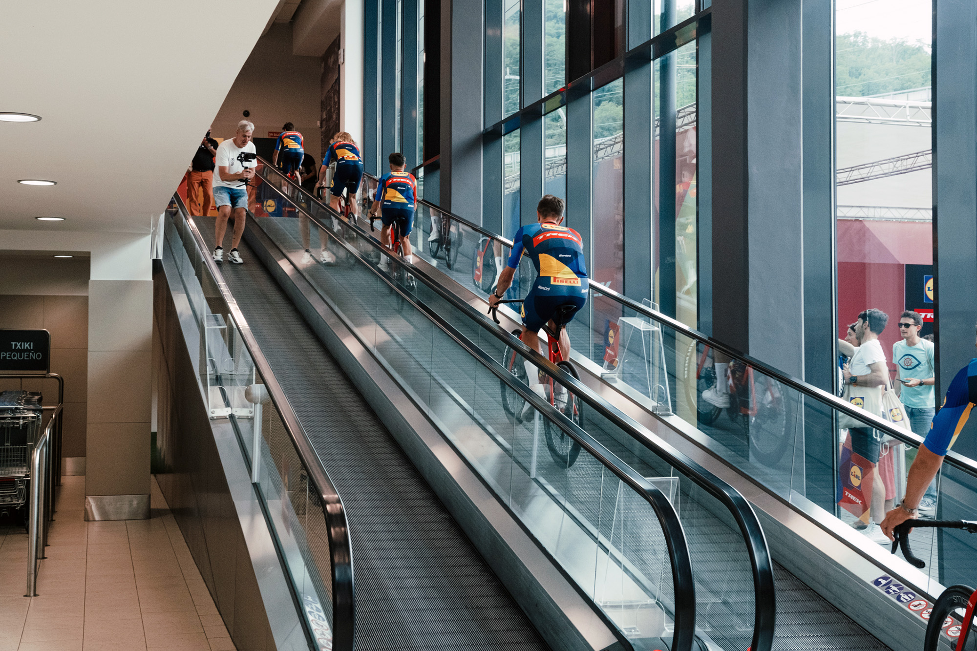 Lidl-Trek riders ride up an escalator in a Lidl supermarket.