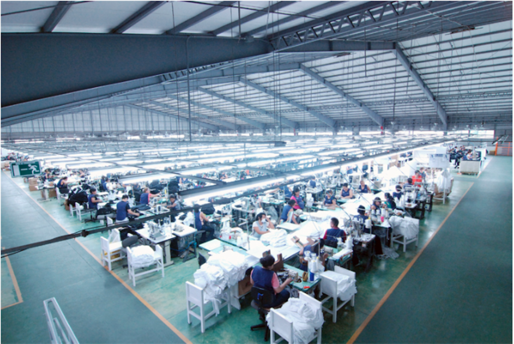 Workers toil on an apparel production line at the former APS El Salvador factory. The factory is clean and well-lit, with orderly rows of stations.