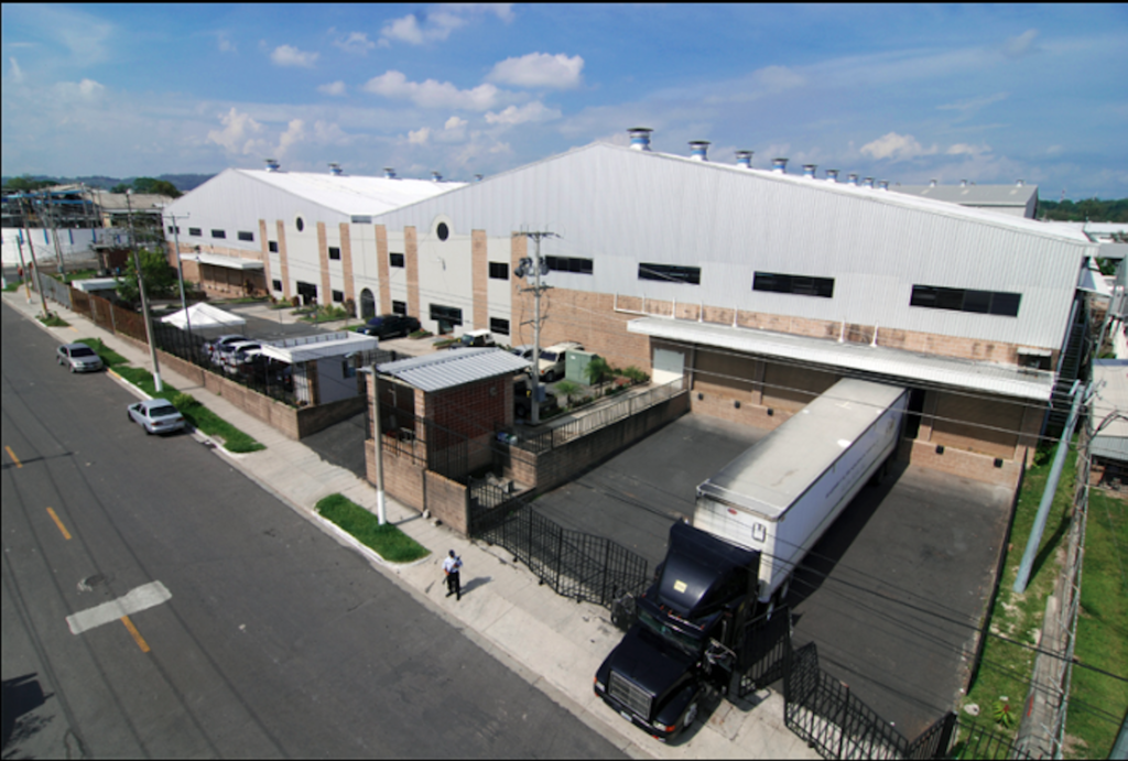 The APS El Salvador factory shown from outside. It's a typical low-multi-storey building, with a large truck and trailer pulled into a shipping by and a security guard out front.