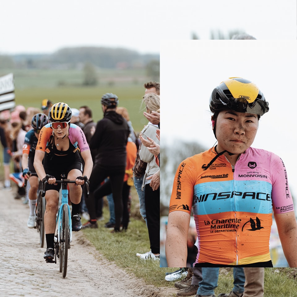 Xin Tang looks impassively at the camera after finishing Paris-Roubaix Femmes, the first male or female Chinese rider ever to do so. Her face and nose especially are coated in dirt, her jersey and arms flecked with grit.