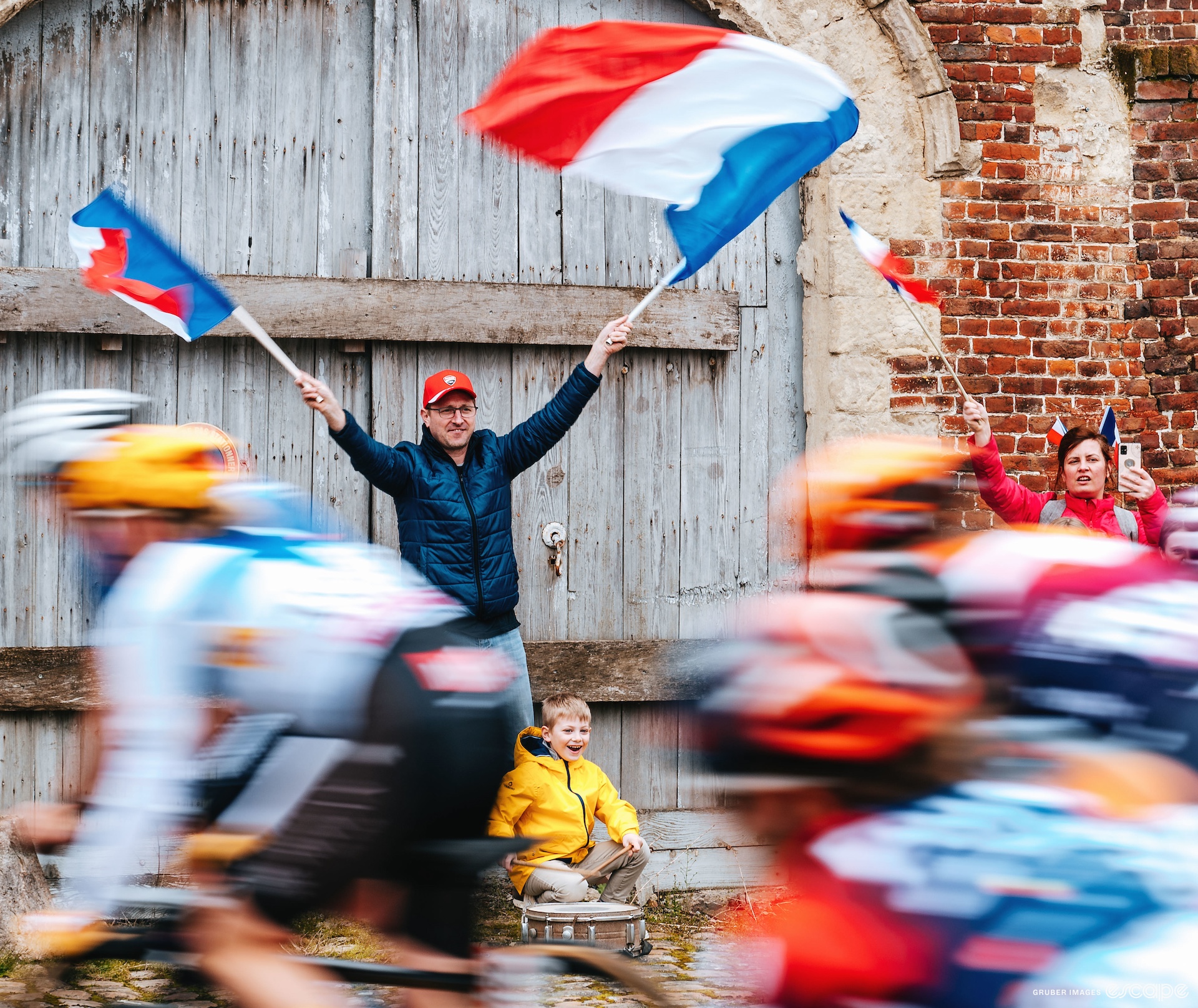 A fan waves French flags at the Paris-Roubaix peloton as they pass in a blur. He stands in front of a large arched door in a red brick building, with his young son at his feet, a delighted look on his face as he plays the drums.