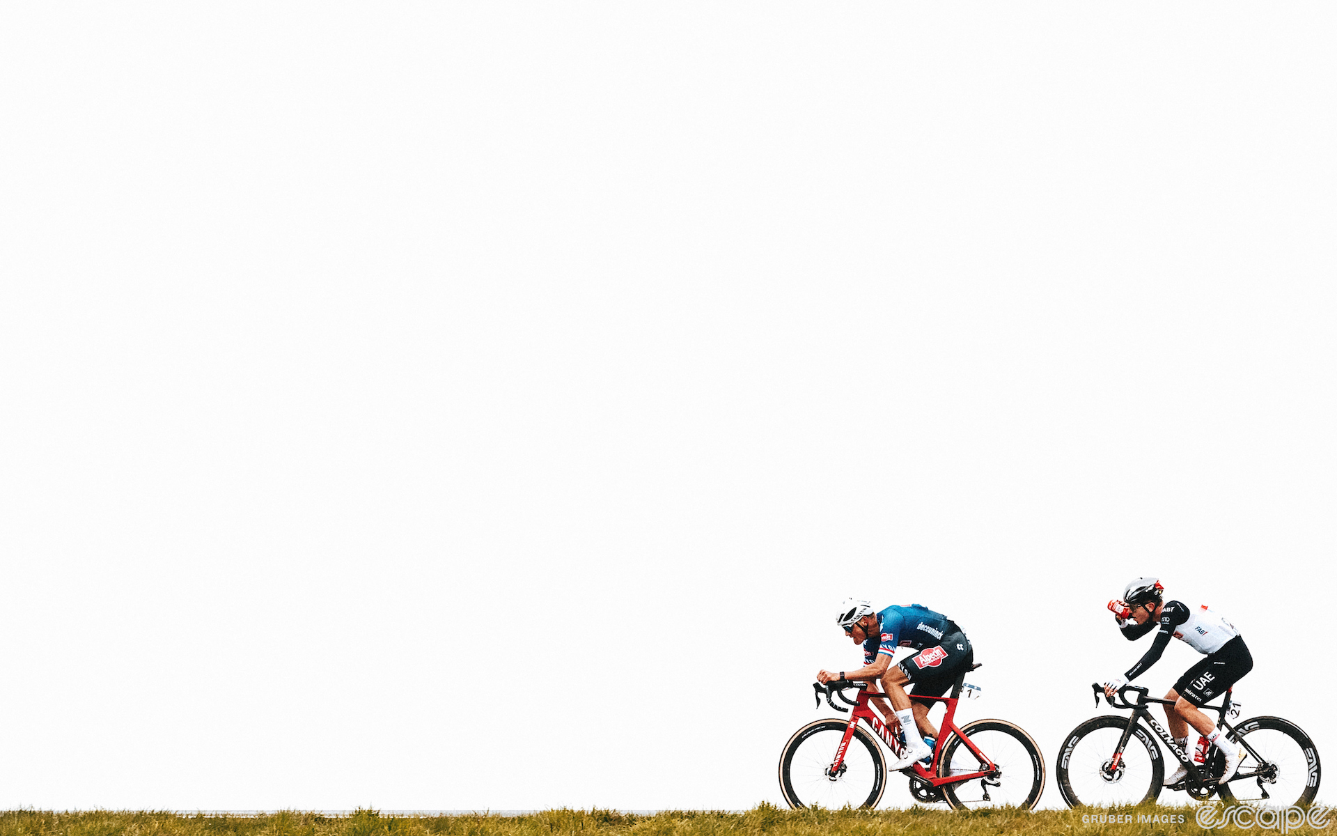 Mathieu van der Poel leads Tadej Pogačar in the 2023 Tour of Flanders. They're the only riders in view, framed from the side with a wide shot. A grassy field just peeks out of the bottom edge of the frame and the sky is blown out in white clouds for a gritty contrast.