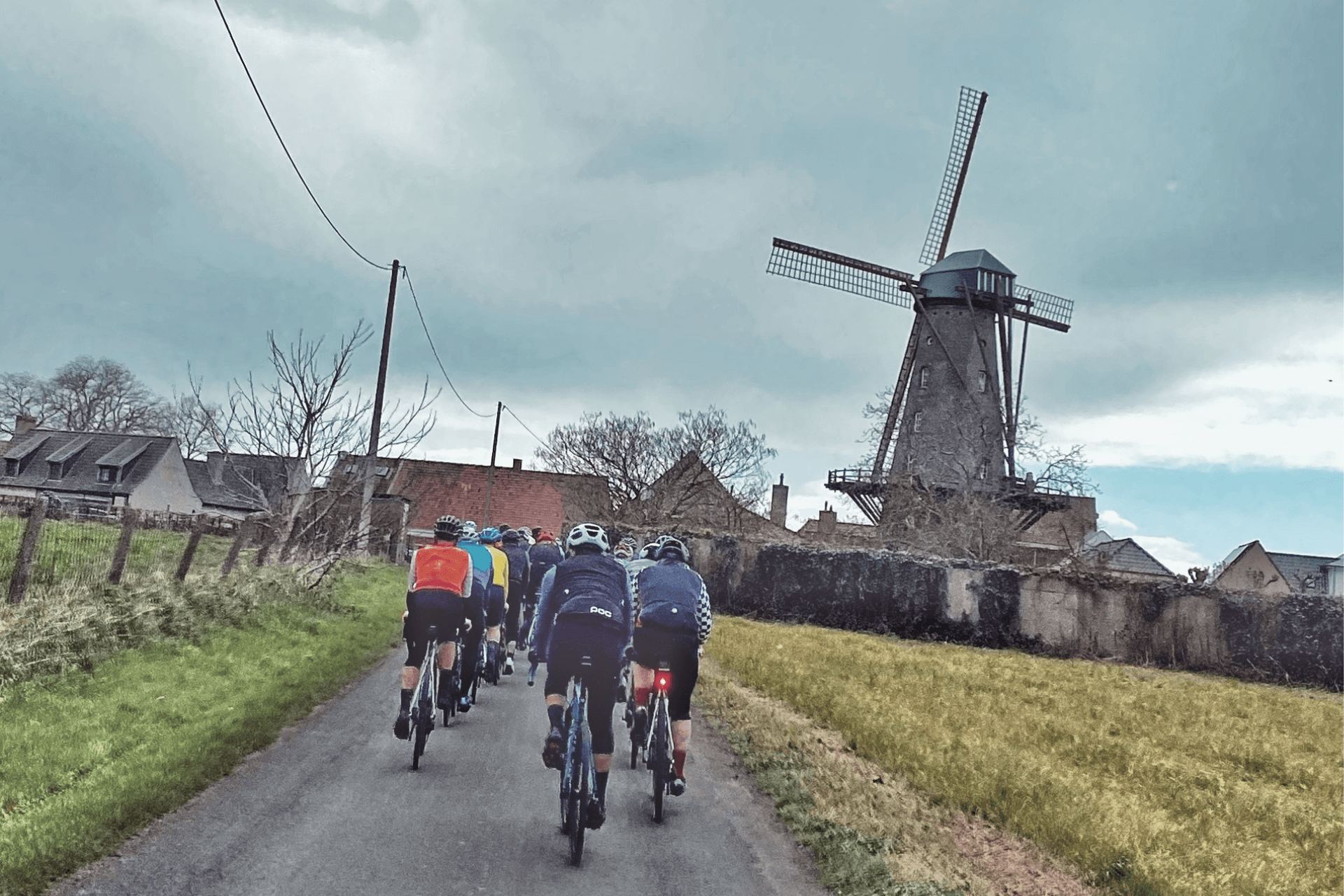 Road cyclists ride two abreast down a narrow farm road. A centuries old windmill features prominently in a small village off in the distance