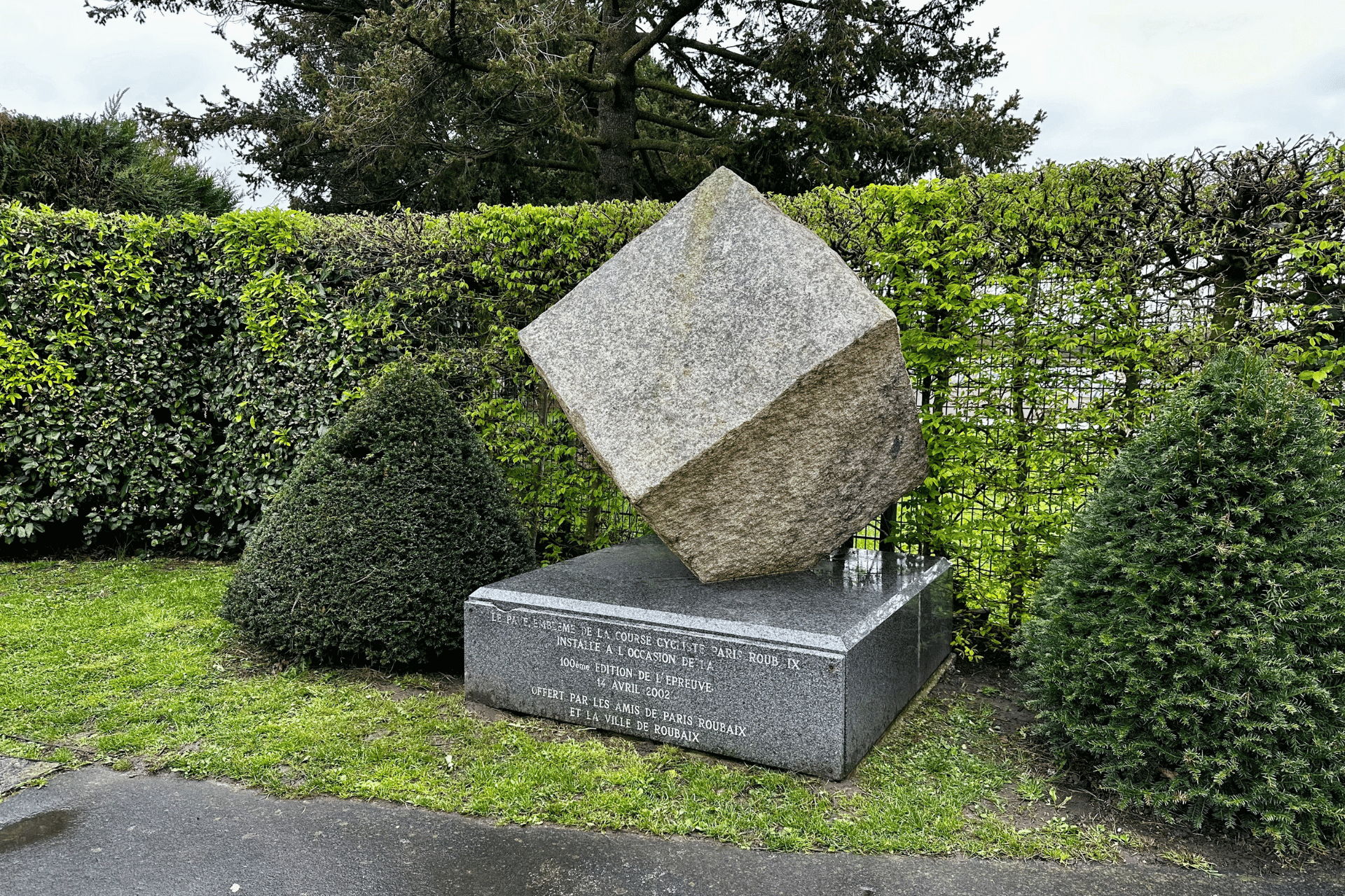A civic monument featuring a large cubic cobblestone. Conical bushes to the left and right frame the monument. The inscription on the plinth reads "The cobble, emblematic of Paris-Roubaix, was installed in commemoration of the 100th edition of the race on April 14th 2002. Made possible by 'The Friends of Paris-Roubaix' and the town of Roubaix" (translated from French)