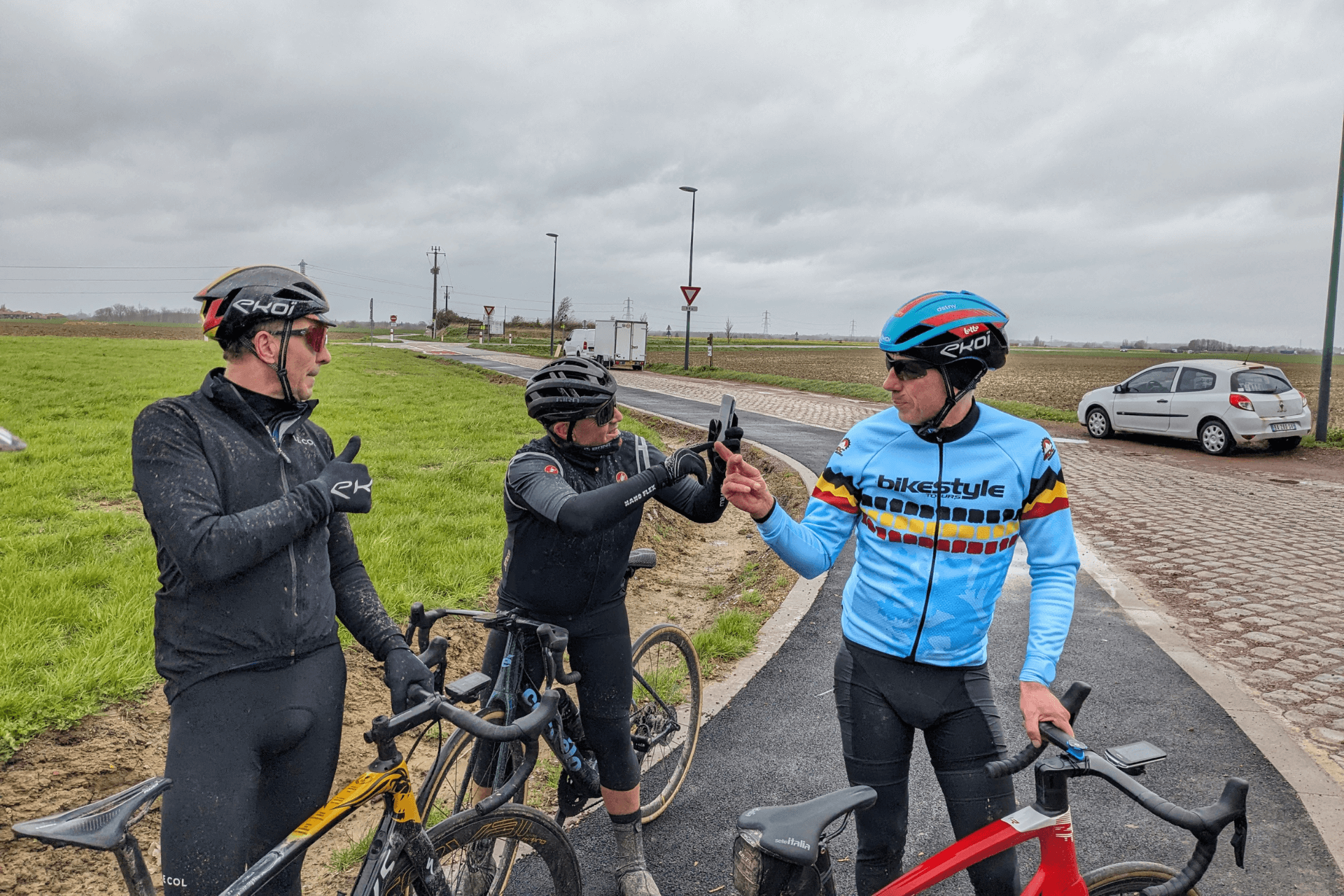 Three stationary cyclists standing on a bike path next to a motorway. They are dressed for cold weather. One is taking a selfie, the other two are engaged in conversation
