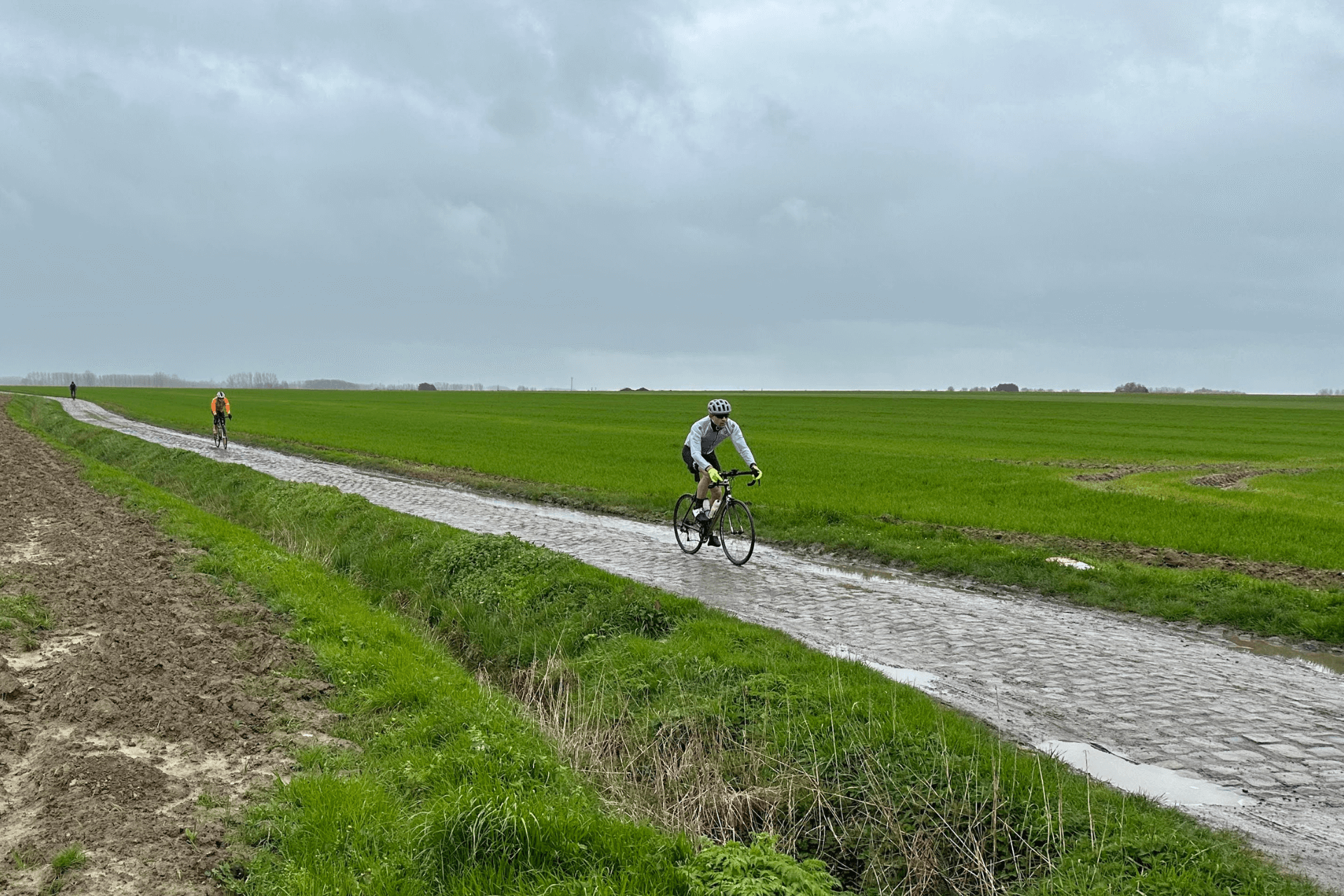 There is a deep green field with a narrow cobbled road in the foreground. Three cyclists, spaced roughly 25 metres apart, ride drop bar road bikes over the cobbles