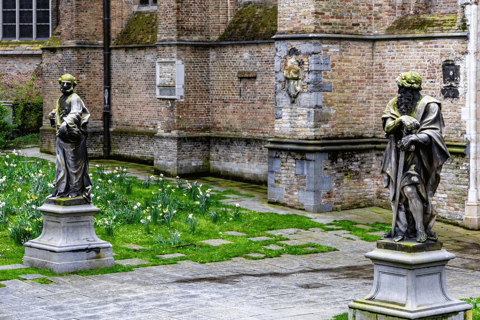 Two moss covered Hellenic-style statues in a moss covered courtyard with the stone walls of a church in the background