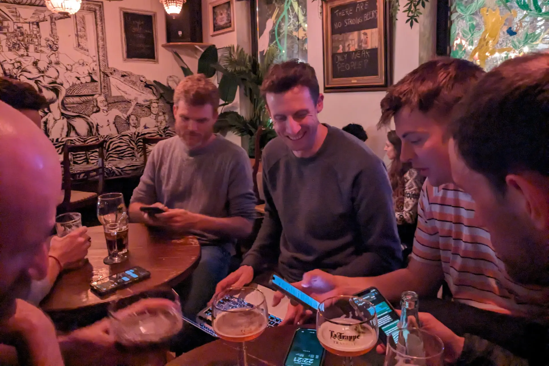 Several people are gathered around two small tables. They are working intently on their phones and laptops and drinking beer