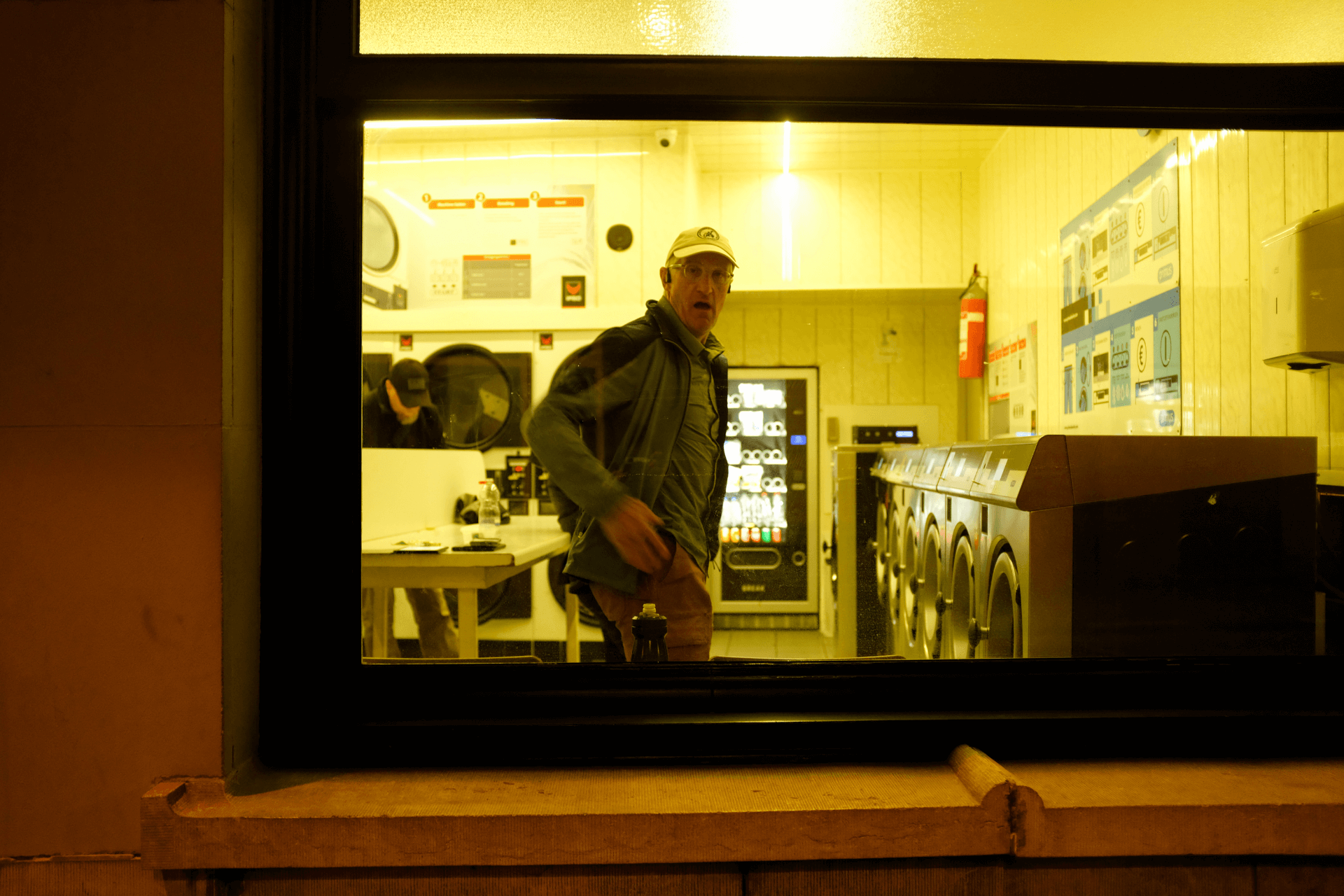 A man with a white cap and casual spring jackets stands in a laundromat, hands in pockets. He is facing out through a window and looking directly at the camera