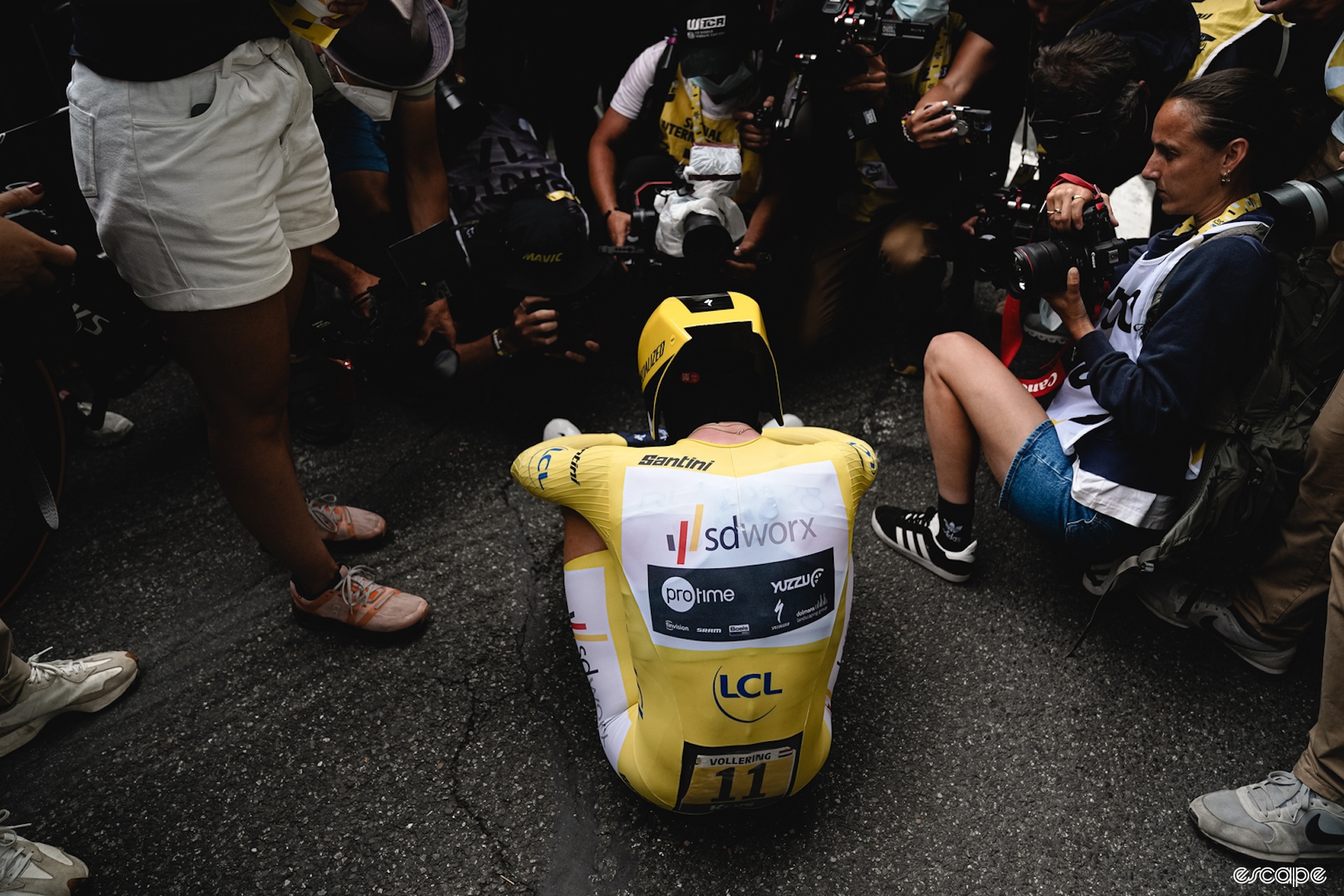 Demi Vollering, wearing a yellow skinsuit, sits on the ground, her back to the camera, surrounded by photographers after winning the Tour de France Femmes in 2023