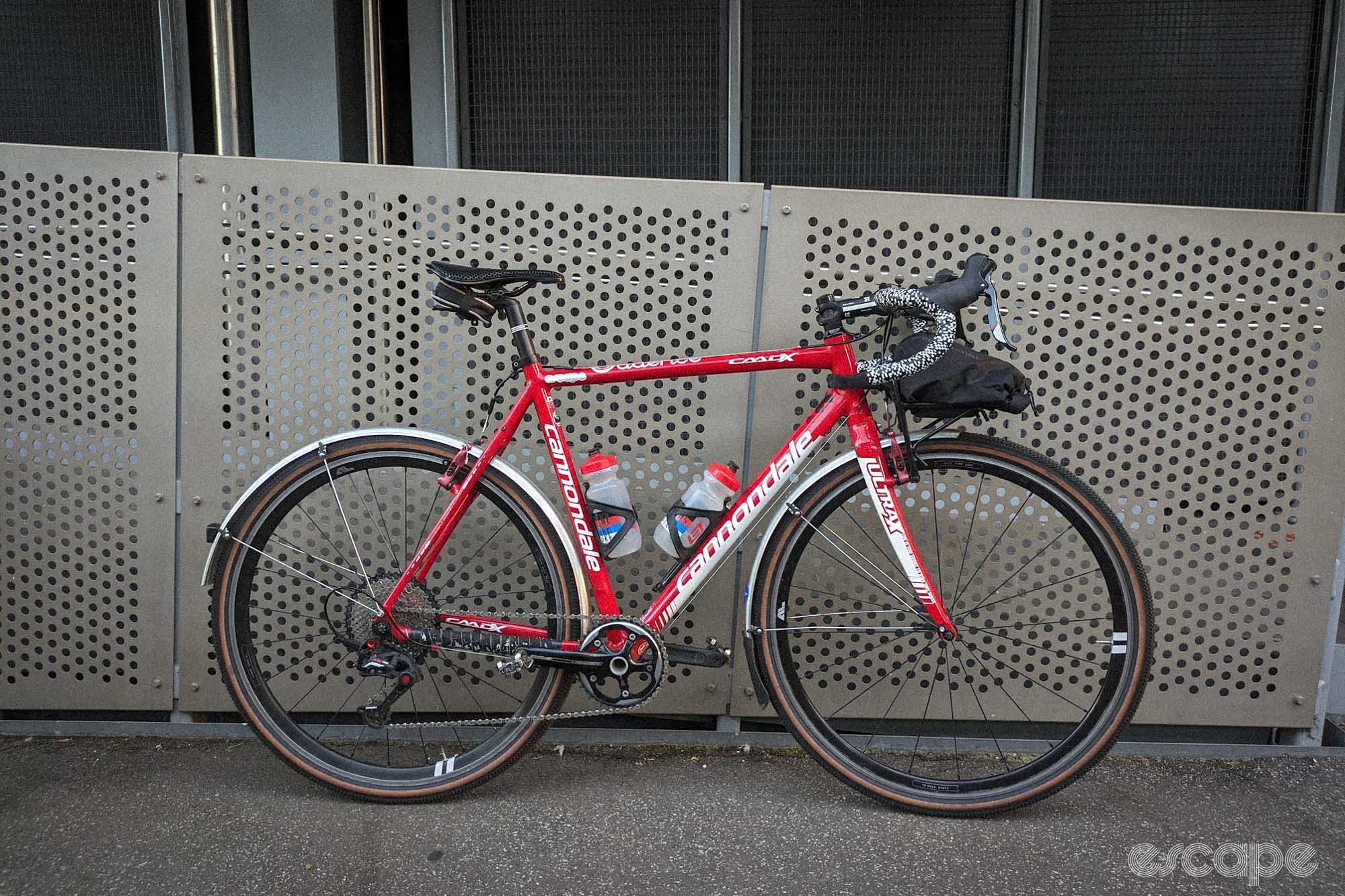 Red Cannondale CAADX, leaning against a fence, with mudguards, fat tanwall tyres, 1x groupset, v-brakes and a front rack with racktop bag. 