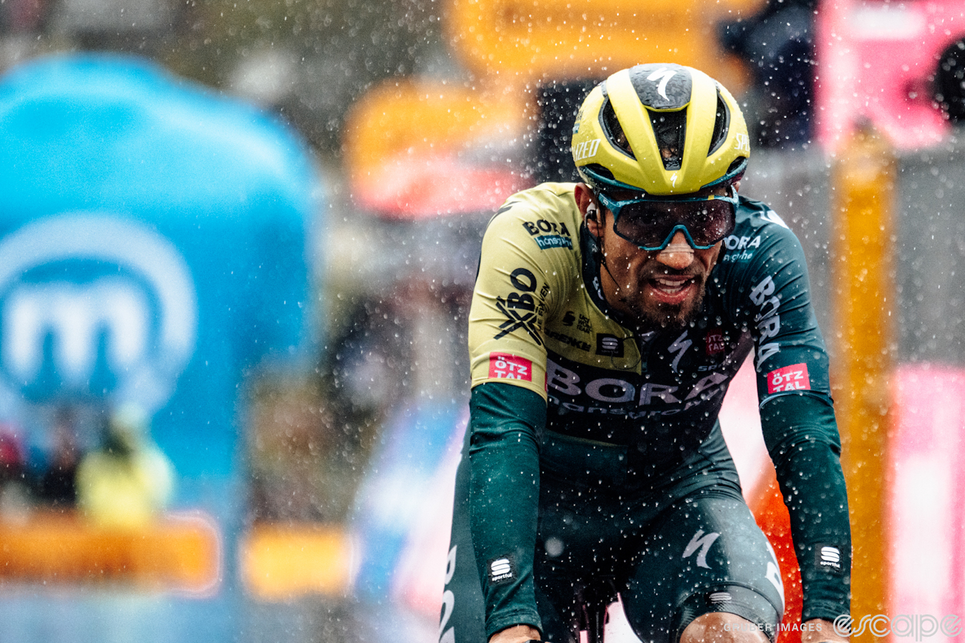Dani Martinez crosses the finish line alone in a rain-soaked stage 16 of the 2024 Giro d'Italia. Road spray and rain droplets are suspended around him and his face is locked in a grimace above his sopping-wet goatee.