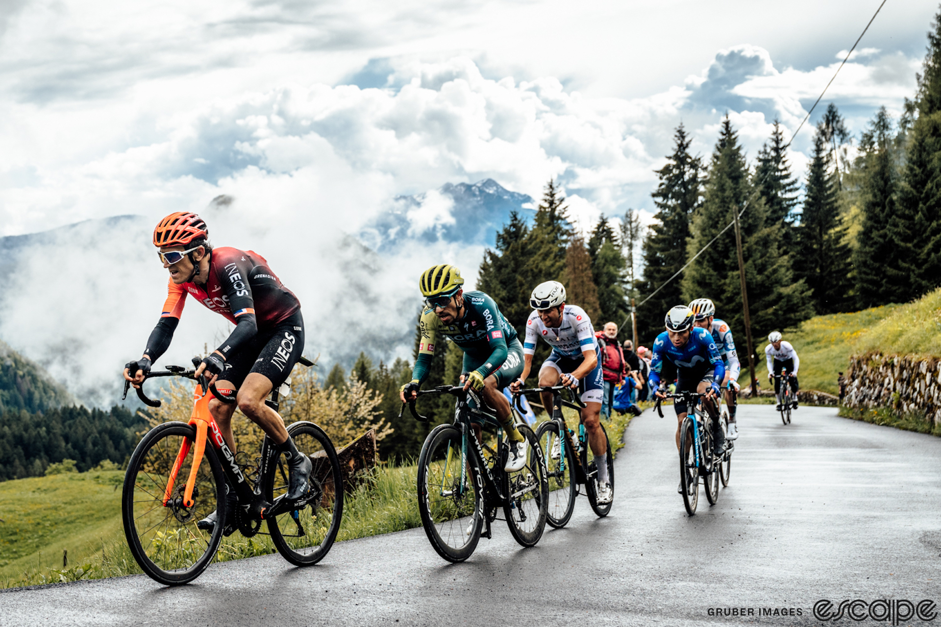 Dani Martinez is locked on Geraint Thomas' wheel as a small group of riders ascends a climb in the 2024 Giro d'Italia. The road is wet, the landscape a lush green, and broken clouds drift over the mountain ridge behind them.