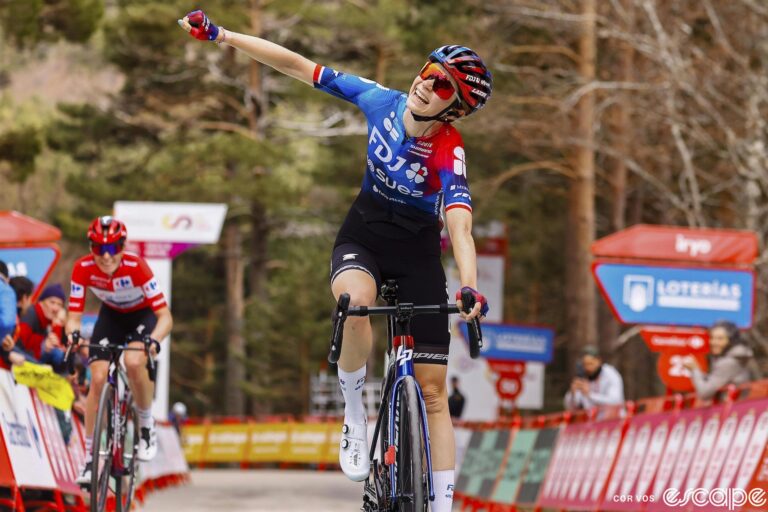 Évita Muzic raises an arm in the air as she crosses the finish line to win stage 6 of La Vuelta España Femenina. Behind, overall leader Demi Vollering is distanced for second place on the day.