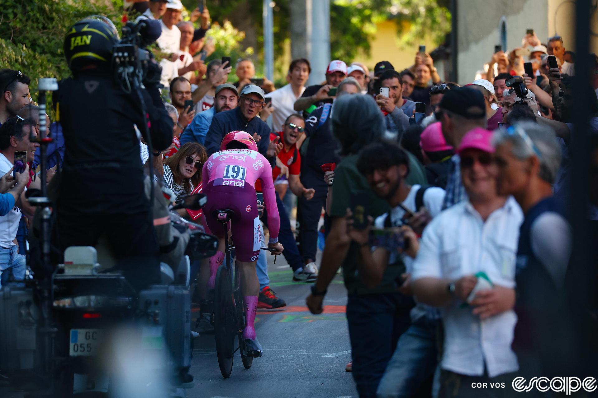 Tadej Pogačar rides the climb to Perugia in stage 7 of the Giro d'Italia. He's alone on his TT bike in his now-allowed pink-and-purple skinsuit, followed by a TV moto as fans lean in to cheer.