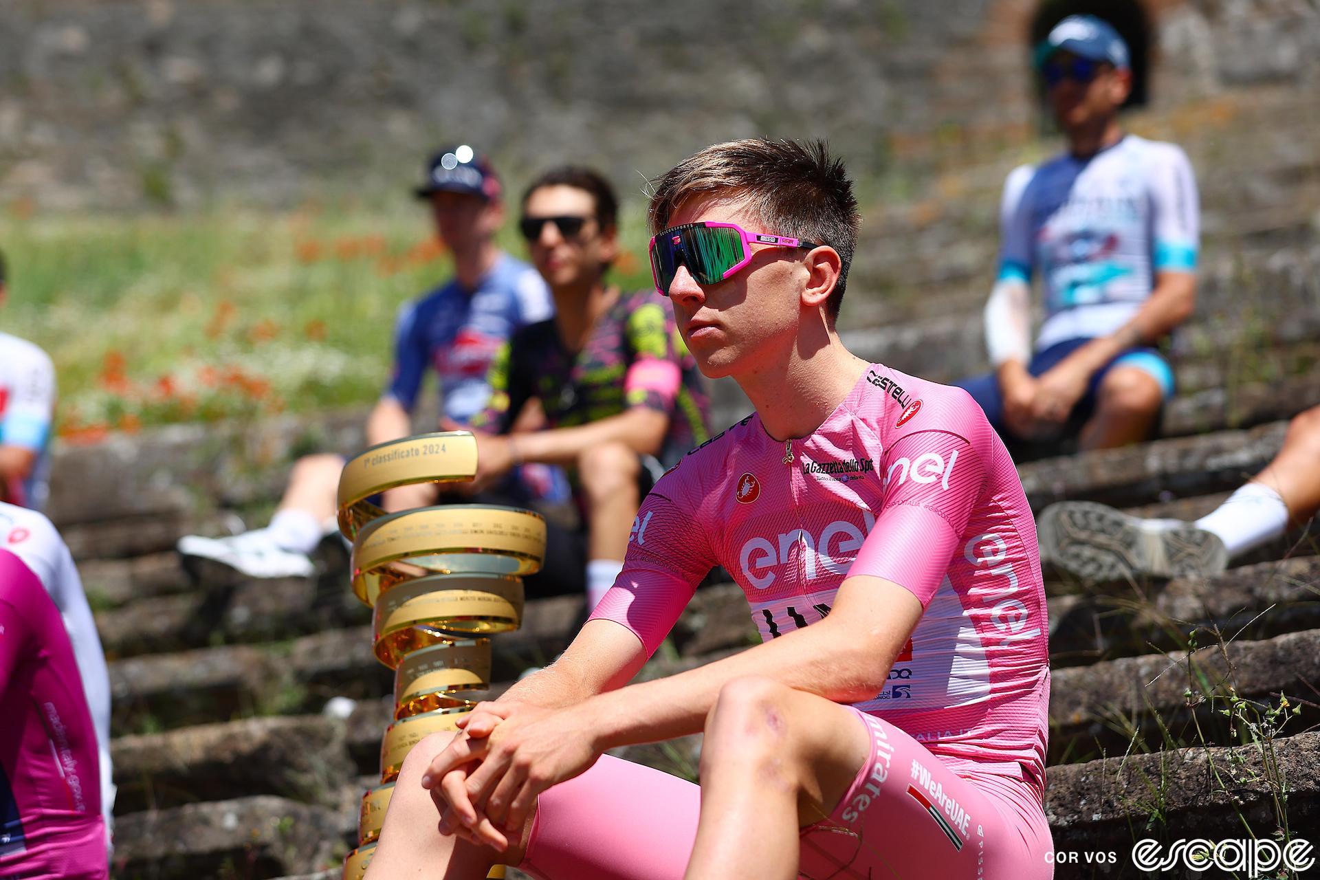 Tadej Pogačar sits on the stone steps of the Anfiteatro di Pompei, clad in a pink race suit of race leader for the Giro d'Italia. Other riders sit near but not next to him. Close by is the spiral trophy for the race's overall winner. He wears large sunglasses that cover most of an otherwise expressionless face.