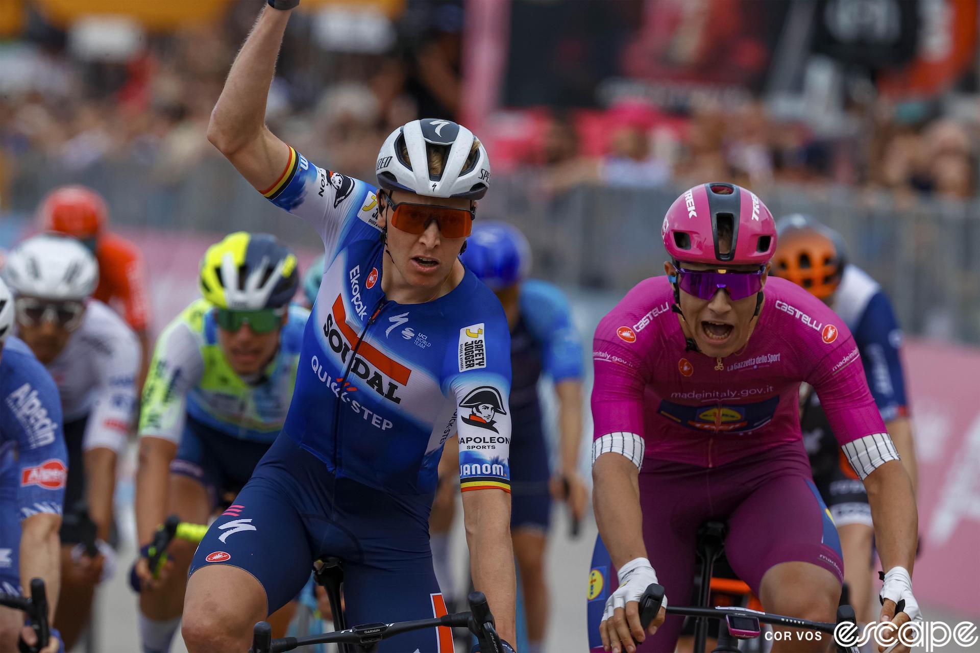 Tim Merlier raises a hand as he crosses the line to win stage 18 of the 2024 Giro d'Italia. To his left, rival Jonathan Milan has his mouth open in a disappointed shout.