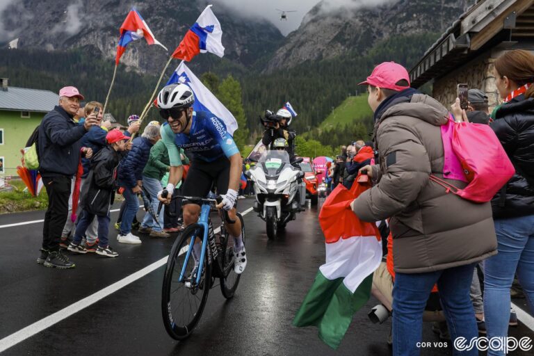 Andrea Vendrame climbs past cheering fans on the way to winning stage 19 of the 2024 Giro d'Italia. The road is wet and the sky behind him dark with rain.