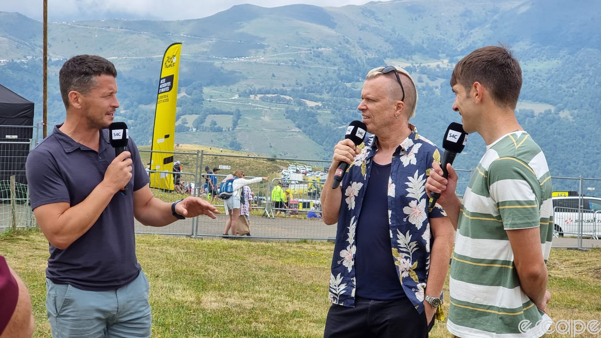 Peredur ap Gwynedd stands on an Alpine hillside at a Tour de France stage with two other men. They're dressed casually, holding TV microphones for the Welsh S4C network.
