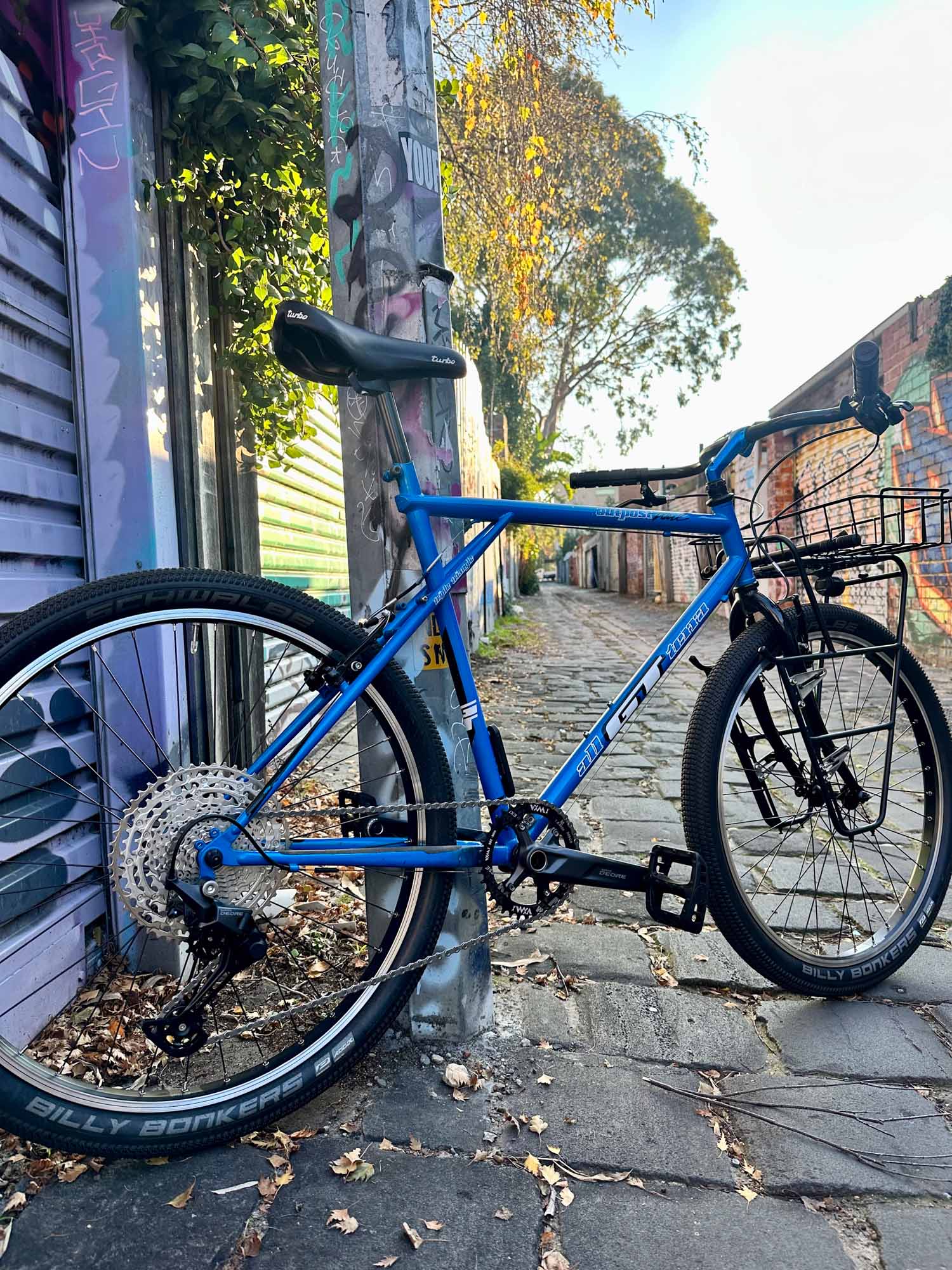 A blue GT mountain bike with swept back bars, a dynamo light and front rack in a cobbled laneway. 