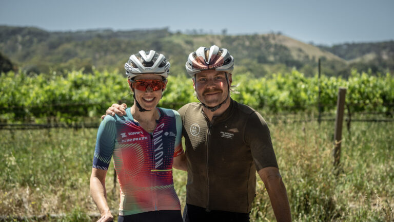 Tiff Cromwell and Valtteri Bottas pose for a portrait. They're in front of a vineyard, wearing cycling kit and helmets, and have their arms around each other.