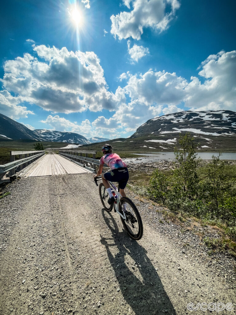 Tiff Cromwell rides a gravel road toward a bridge in the Finnish wilderness. Rounded peaks dappled with snowfields loom in the distance under a sky speckled with clouds, and a bright sun overhead.
