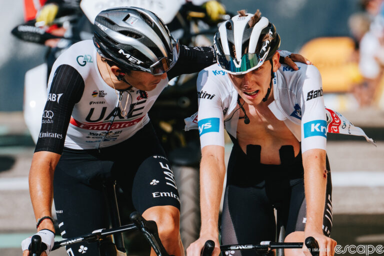 Tadej Pogačar crosses the finish line on stage 17 of the 2023 Tour de France. He has a hollow, vacant expression and his jersey is unzipped as teammate Marc Soler lays an arm across his shoulders in consolation.
