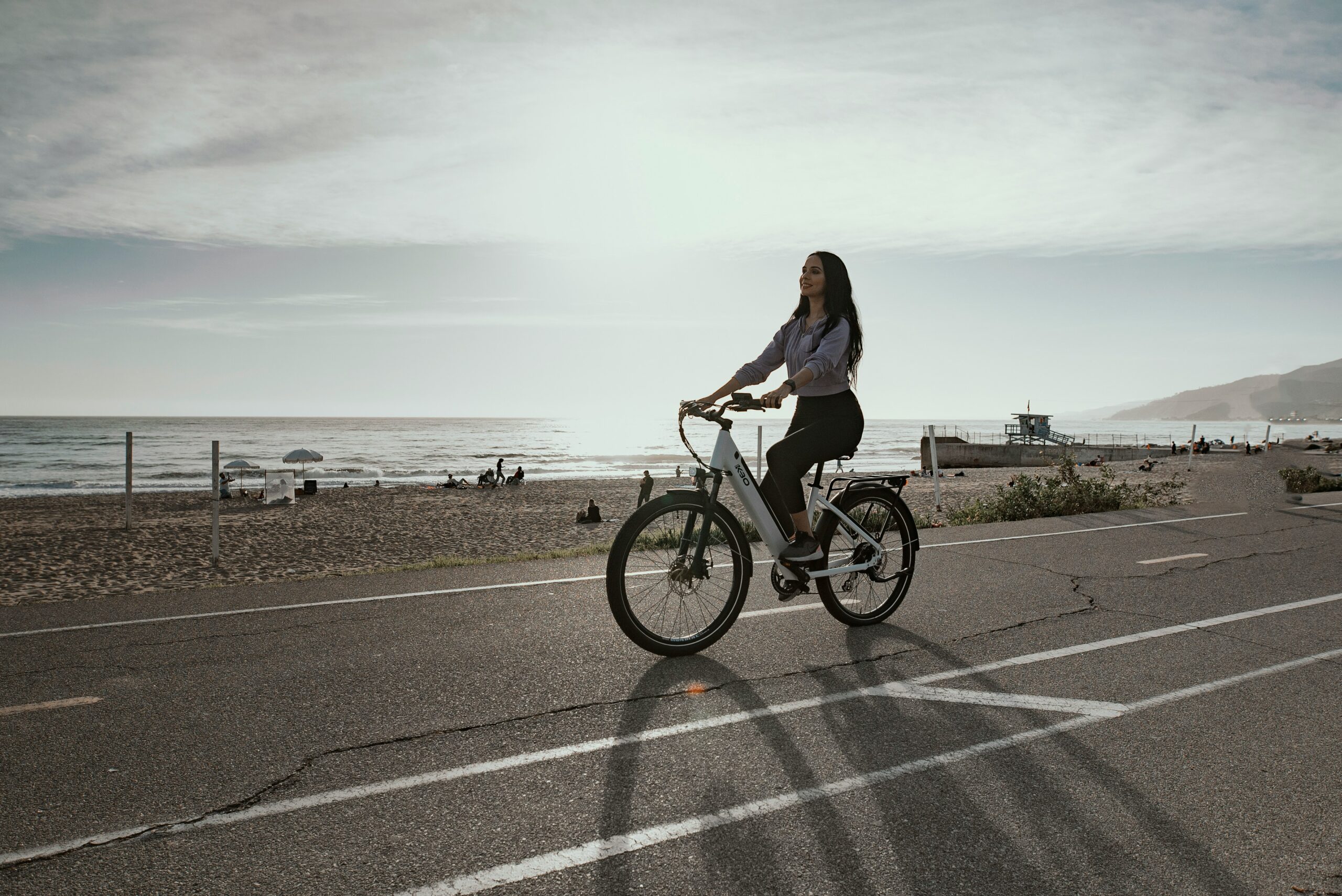 A woman rides an e-bike on a path next to the beach, with the sun behind her.