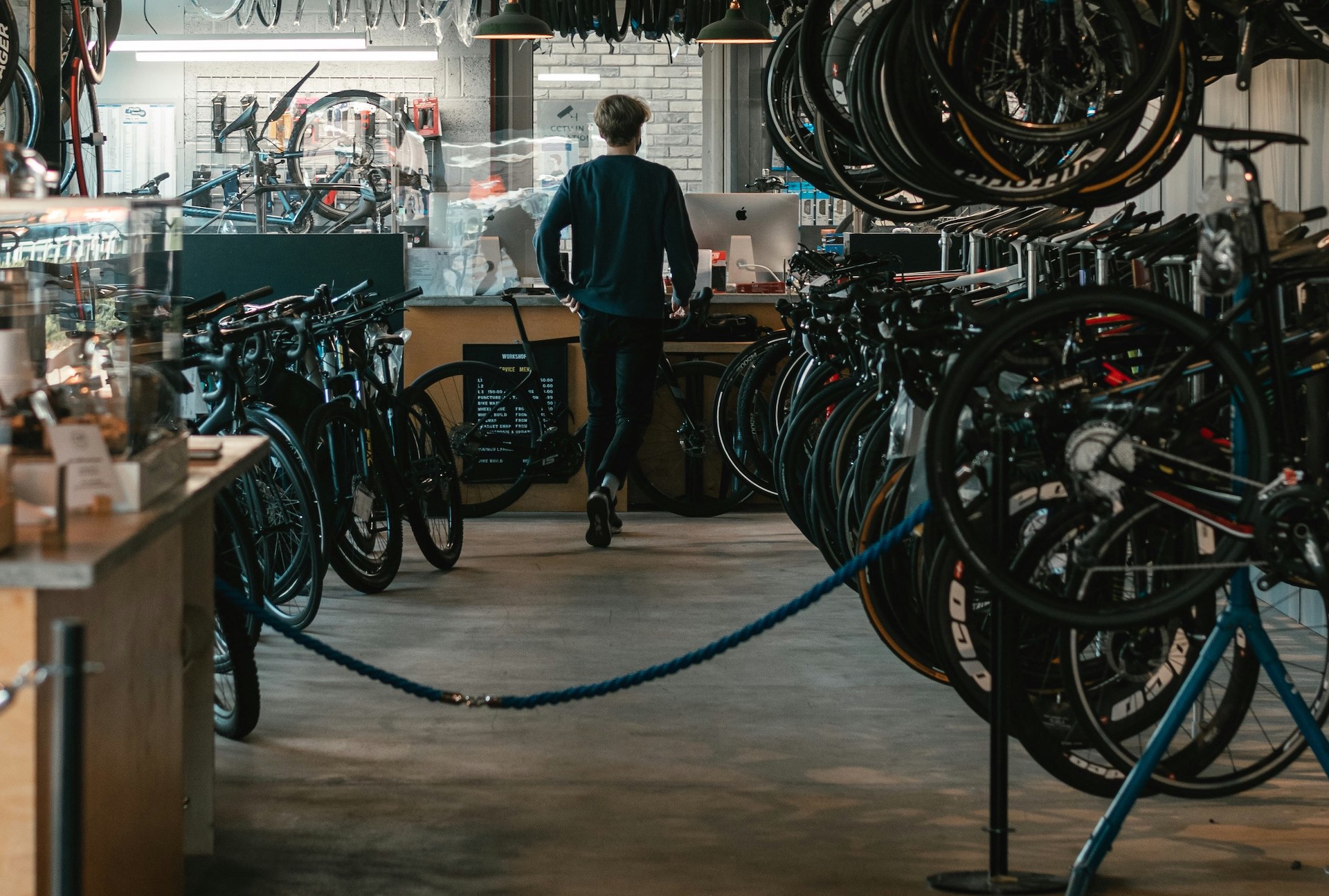 A man walks away from the camera down an aisle in a bike shop, lined by two rows packed with bikes. In front of him is the workshop area.