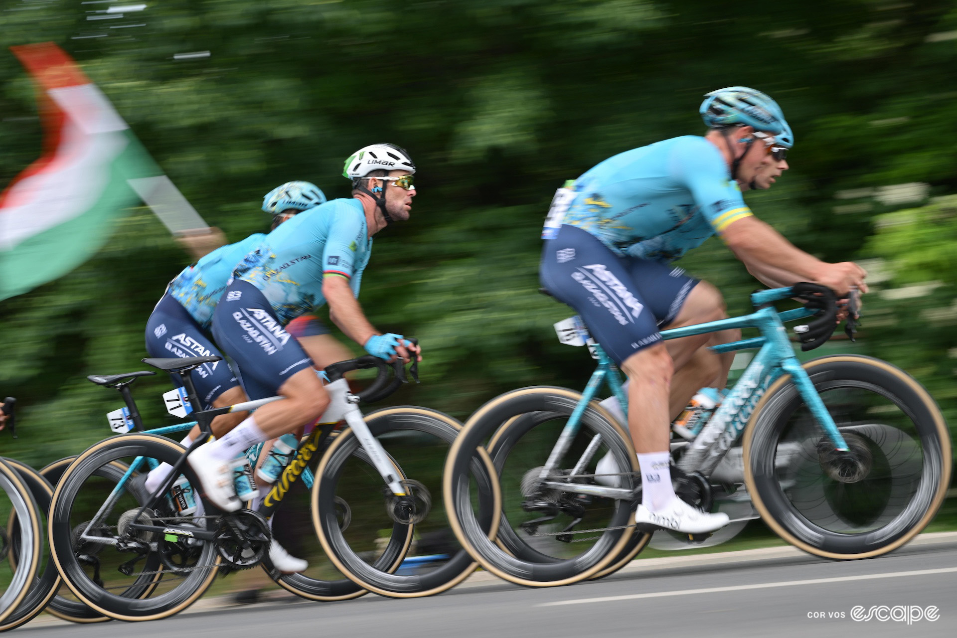 A side view of Mark Cavendish riding among his Astana Qazaqstan teammates during the Tour of Hungary, an out of focus Hungarian flag flying in the background.