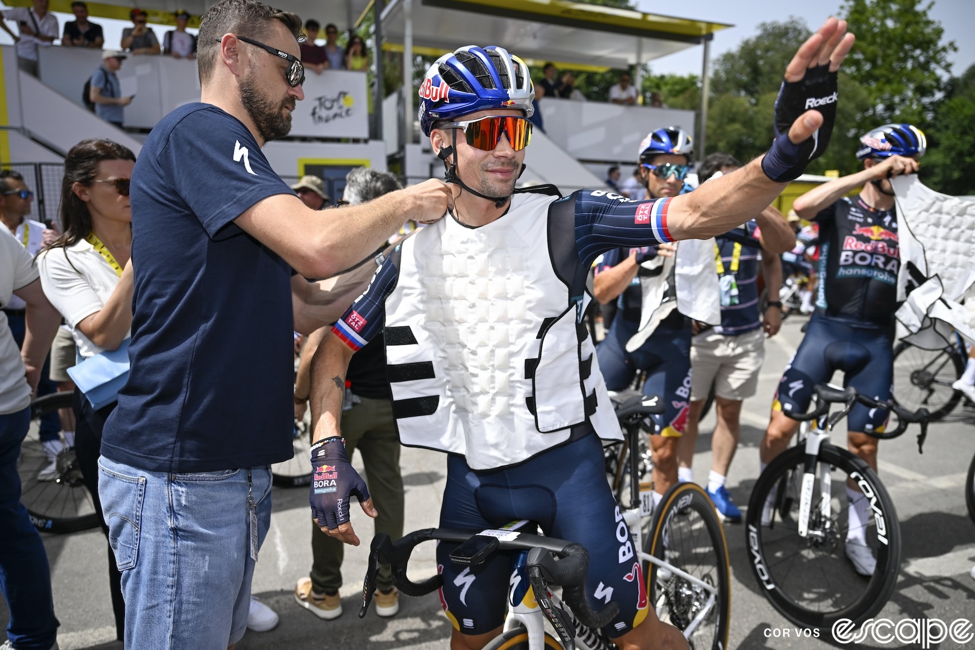 Primož Roglič waves to the crowd as a team staffer fastens a white cooling vest on his torso.