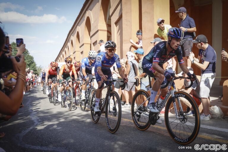 Primož Roglič leads Remco Evenepoel and a select group of chasers on the Côte di San Luca climb on stage 2 of the 2024 Tour de France. They're racing past a long, two-story building of red sandstone with many arches, the road lined with fans, and other riders including Egan Bernal and Carlos Rodriguez behind.