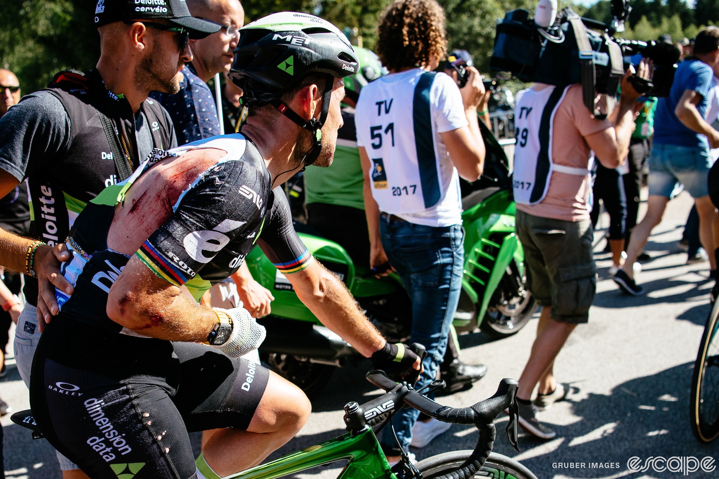 Mark Cavendish rides slowly after the finish of stage 4 of the 2017 Tour de France. He's assisted by a Dimension Data soigneur, and cradles his right arm close to his chest. His jersey is badly torn and his right arm and shoulder are scraped and bloody.
