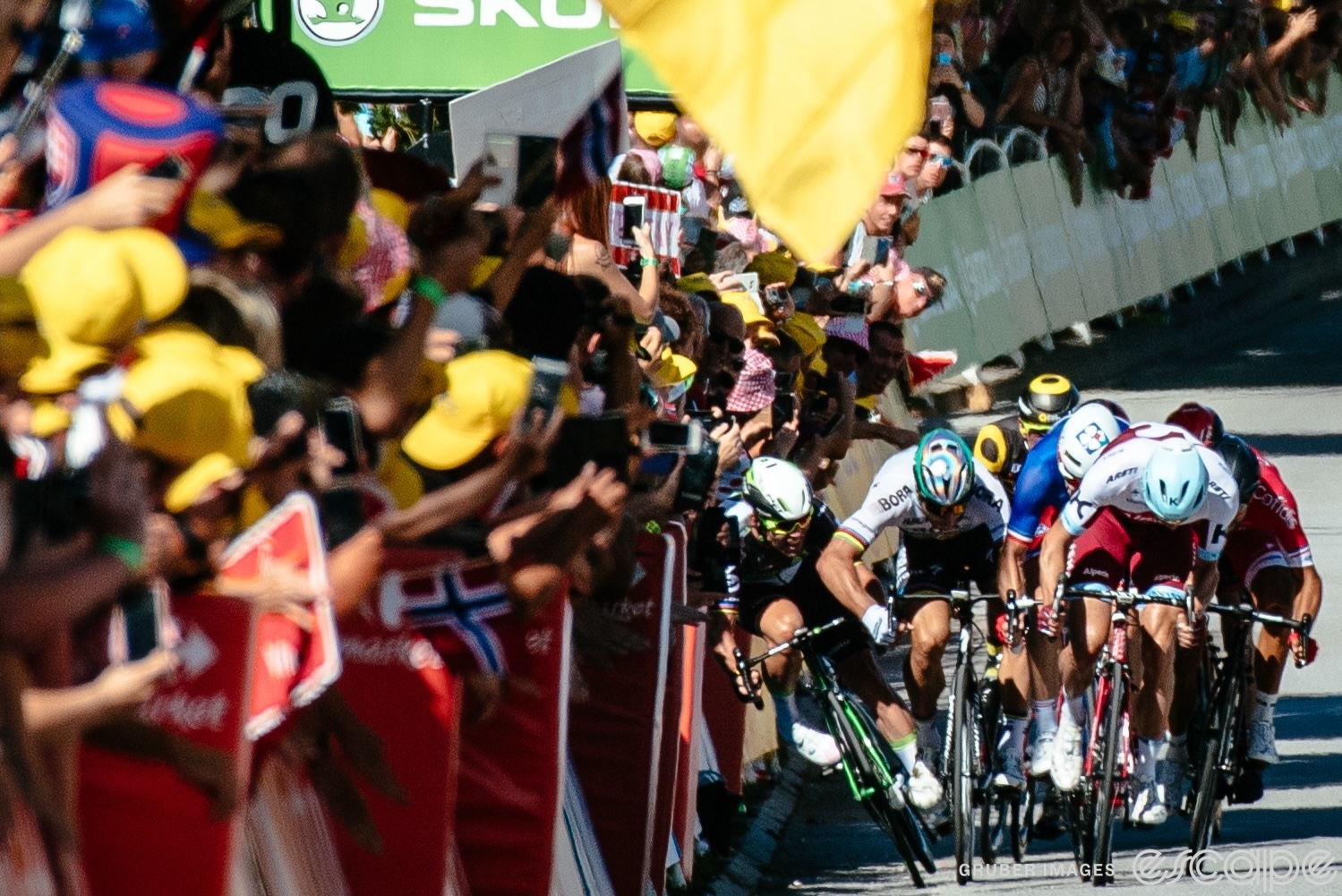 Riders accelerate for a sprint on stage 4 of the 2017 Tour de France. Their heads are down, and Peter Sagan's right elbow juts out as he makes contact with Mark Cavendish. Cavendish is squeezed against the barriers and is falling but has not yet hit the ground.