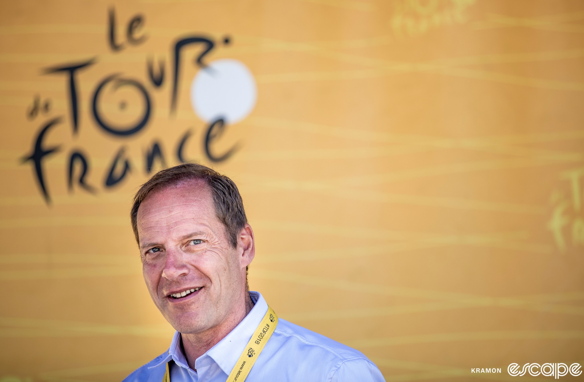 Christian Prudhomme looks at the camera with a slight smile on his face. He stands in front of a large banner with the Tour de France logo.