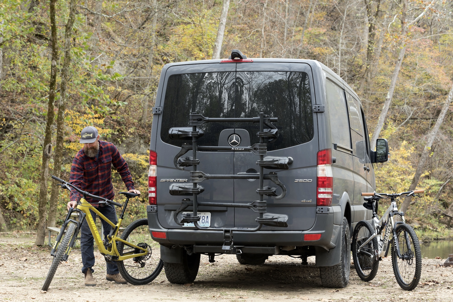 A bearded man unloads a mountain bike next to a Sprinter-style van with a Küat Transfer v2 rack on the back.