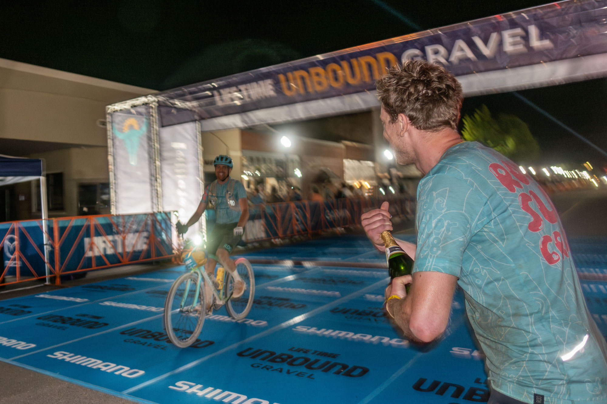 The last of the beach cruiser trio crosses the line after dark, smiling, while one of his teammates pops a bottle of champagne.
