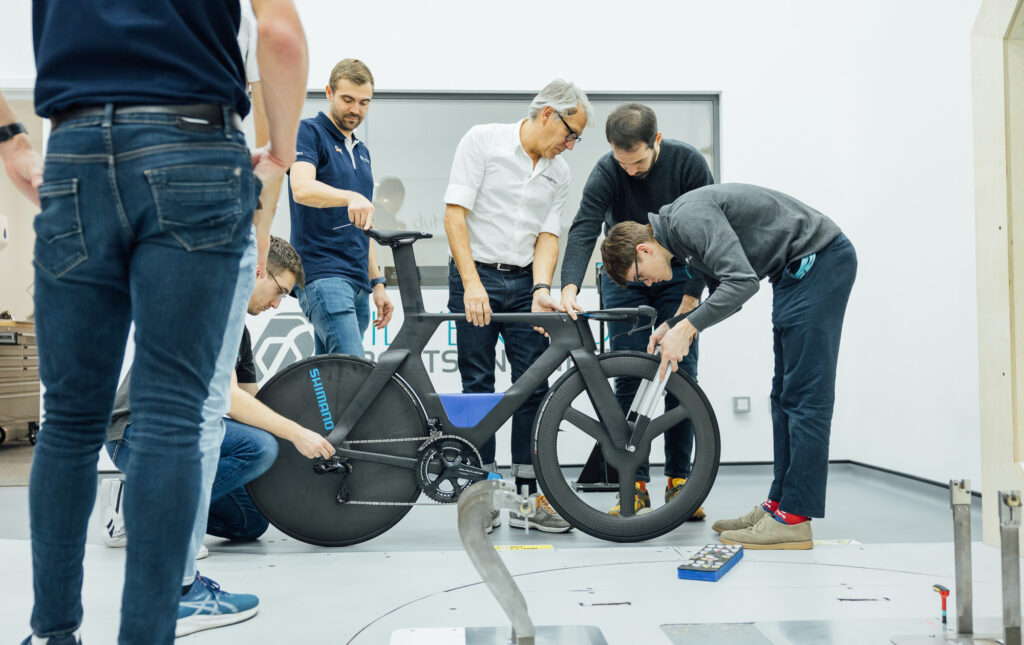 The image shows seven people setting up the new bike in the wind tunnel.